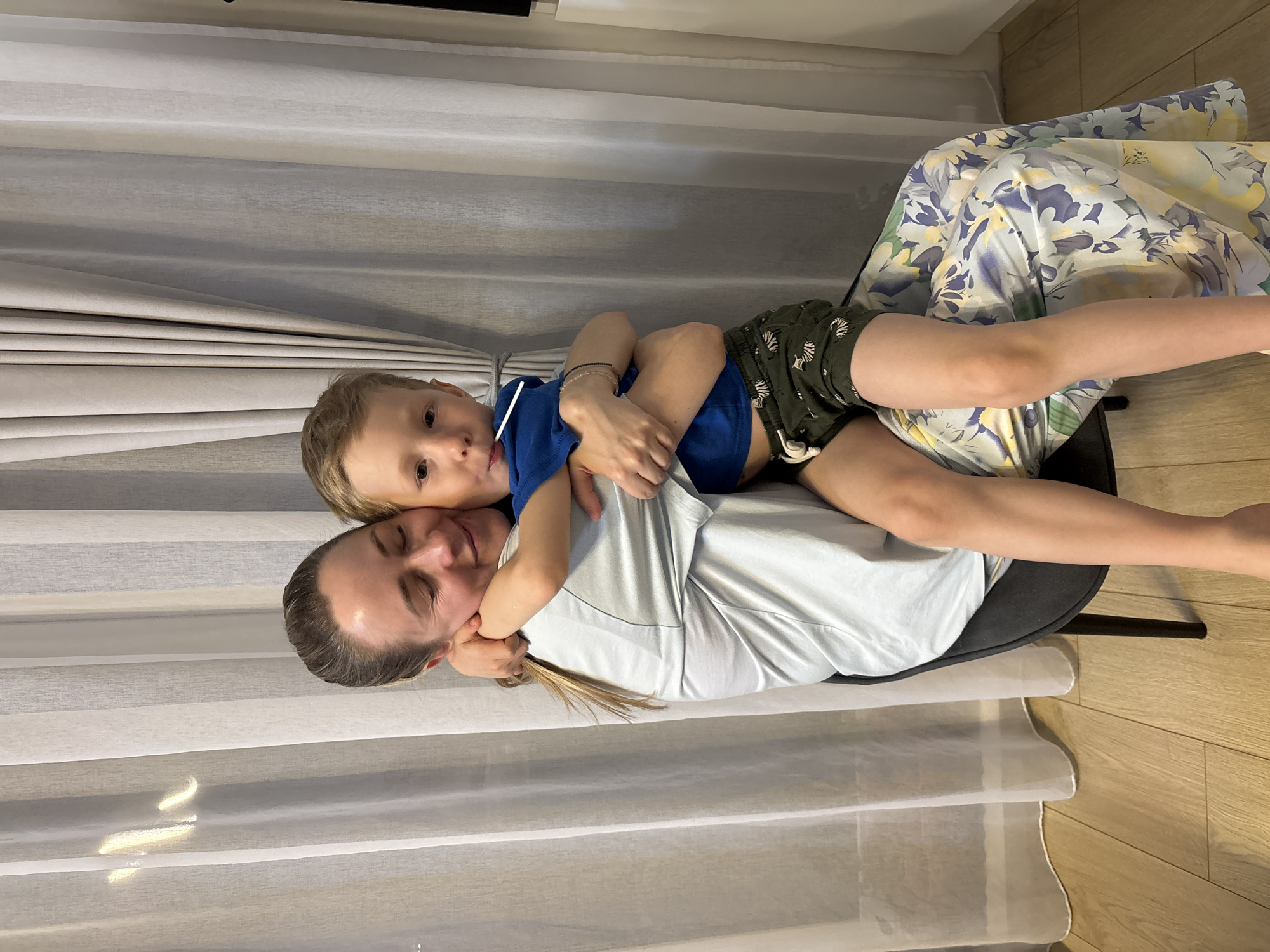 A woman and a young child are embracing while sitting on a chair. The woman is wearing a light-colored shirt and has her hair pulled back. The child is wearing a blue shirt and dark shorts with a zebra pattern. They are indoors, with light-colored curtains in the background and a wooden floor. The chair is covered with a floral patterned cloth.
It is not possible to verify if the image was taken in Chaikovsky, Russia.