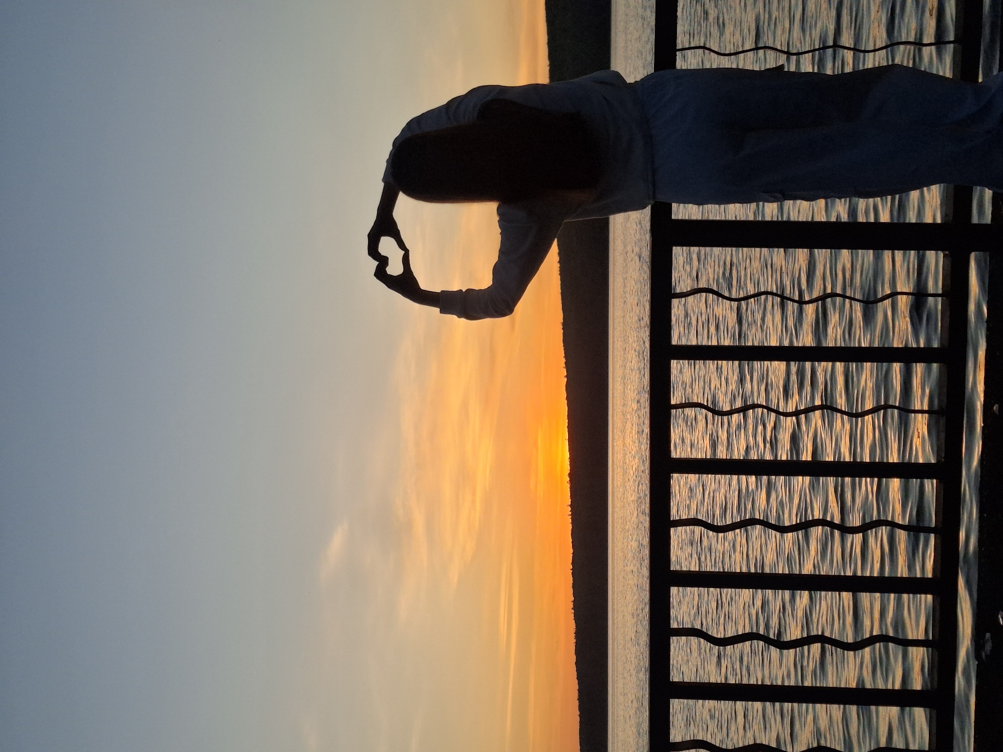 Here's a detailed description of the image:
The image depicts a person standing on a balcony overlooking a body of water during sunset. The person is silhouetted and appears to be making a heart shape with their hands. The sunset creates a vibrant orange and yellow hue on the water and sky, and the balcony railing is visible in the foreground. The location is the city of Chaikovsky, Russia.