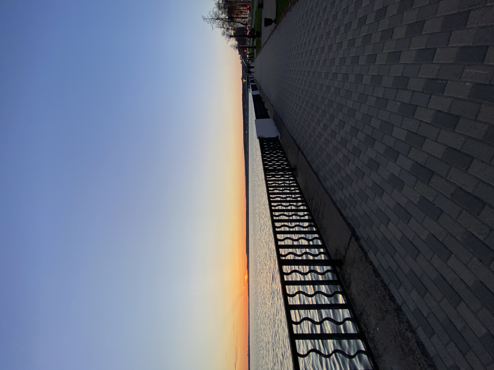 The image depicts a waterfront scene in Votkinsk, Russia. The photograph is taken from a high angle, looking along a paved brick pathway. To the right of the pathway is a metal railing with a wavy design, separating the path from the water. The water surface is visible behind the railing, reflecting the colors of the sunset or sunrise. The sky transitions from blue to orange and yellow near the horizon, indicating either dawn or dusk. Trees and street lights can be seen in the distance. The photograph captures the transition of day into night, or vice versa.