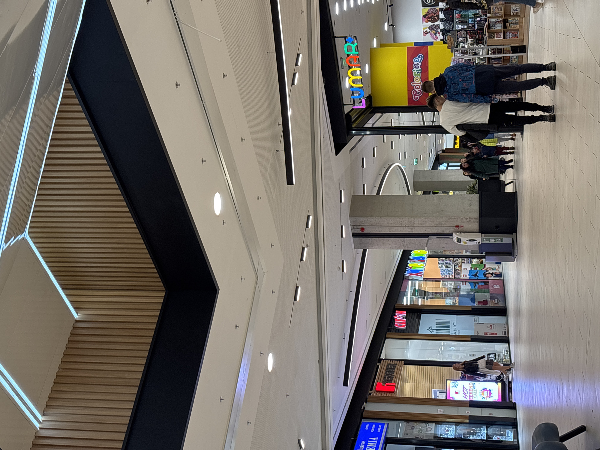 This indoor shot depicts a shopping mall, presumably in Sofia, Bulgaria, showcasing its interior architecture and retail stores. The image captures a high perspective within the mall. On the right side, several individuals are walking, seemingly shopping. Their movements are captured in a general, non-specific manner.
Retail shops are visible with storefront displays, including signs such as "Coloring", "Baker", "ANIC", "BERGNER", "EMIA". These stores display a range of merchandise. Architectural details include a textured ceiling with recessed lighting, a unique stairwell with contrasting black and light-colored wooden elements, and structural columns. The mall has a modern design, and its lighting highlights the retail spaces. The focus of the image remains on the interior architecture and the shopping environment.