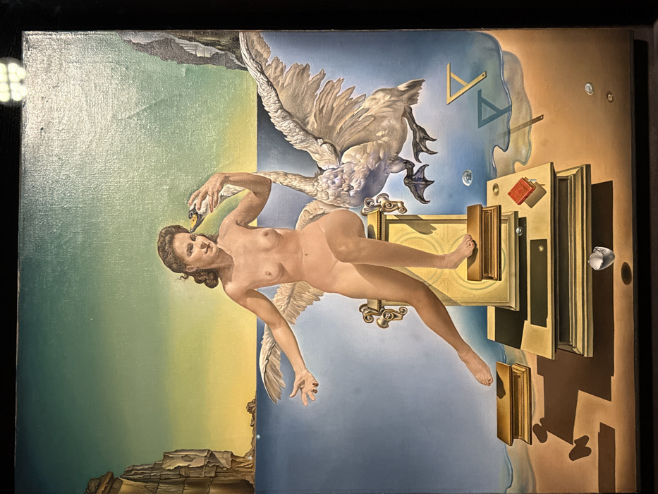 The image displays a surrealist oil painting, rendered with a dreamlike quality and rich detail. The painting is oriented 90 degrees clockwise in the provided image, but it depicts a scene as if viewed upright.
At the center of the composition is a nude female figure, gracefully suspended or floating. She has dark, curly hair and a serene expression on her face. Her body is rendered with soft, realistic flesh tones. Her right arm is raised, and her hand gently touches the head of a large white swan. The swan's wings are spread wide, as if in mid-flight or descent, and its webbed feet are visible. The swan's head is tilted towards the woman, its beak slightly agape. The woman's left arm extends downwards, with her hand open, while her legs are bent, her right foot seemingly interacting with a golden, ornate frame.
The background of the painting is divided into several distinct, yet fluid, sections. On the left side, there is a landscape of rugged, rocky cliffs or formations, rendered in shades of green and yellowish-brown, suggesting land meeting water or sky. This transitions into a brighter yellow-green area that forms a horizon line. On the right side, the upper portion features a light blue sky or ethereal space, which gradually blends into a sandy or brownish-orange ground plane towards the far right.
Scattered throughout the right side of the painting are various surreal elements. Numerous wooden frames, some appearing like empty picture frames and others resembling doors or portals, are depicted as if falling or suspended in space. These frames cast long, stark shadows, suggesting a strong, dramatic light source from the upper left. Several books are also present; a small, vivid red-covered book is visible within one of the frames, and another book with a prominent gold spine is positioned such that the woman's right foot appears to be pressing into it, or it is emerging from the frame beneath her foot. Other open books are shown throughout the scene, some appearing to float.
In the blue sky section, faint outlines of geometric shapes, specifically triangles, are visible, adding to the abstract and intellectual undertones of the piece. Several translucent, spherical drops or pearls are scattered across the blue and brownish sections of the background. Near the bottom right, a small, white, fractured object, possibly resembling a broken eggshell or a piece of statuary, lies on the ground plane.
The overall impression is one of a symbolic and mythological narrative, perhaps referencing the classical myth of Leda and the Swan, reinterpreted through a surrealist lens. The juxtaposition of the realistic figures with the impossible, floating objects and fragmented architecture creates a dreamlike, enigmatic atmosphere. The painting exhibits a meticulous technique, characteristic of surrealist masters, with strong contrasts in light and shadow and a rich, symbolic color palette. No discernible text is visible within the image.