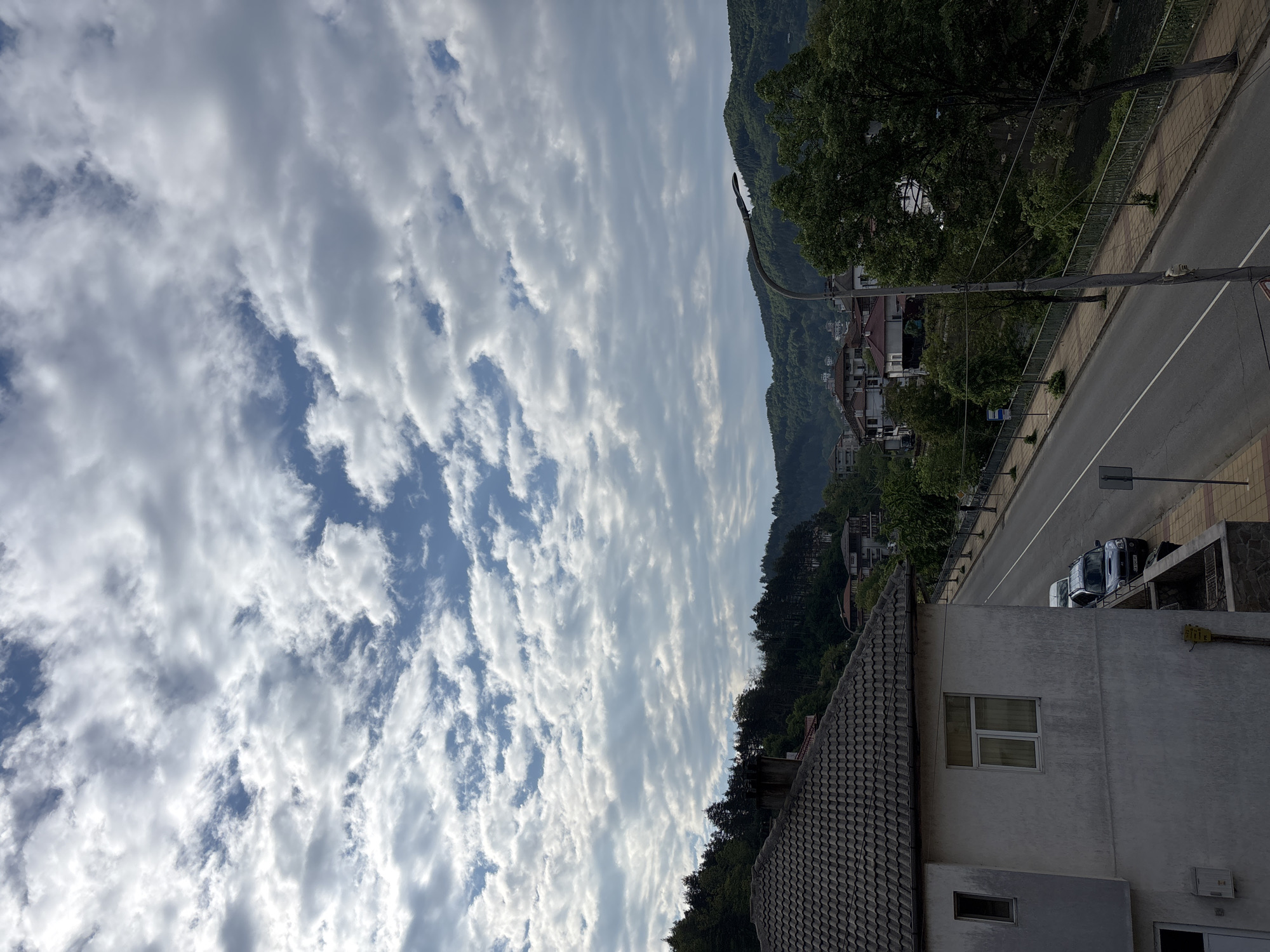 The image shows a daytime scene in Zlatograd, Bulgaria, featuring a street with a mountain backdrop and a cloudy sky. The sky is filled with rows of altocumulus clouds, revealing patches of blue. A road runs along the right edge of the frame, lined with a metal fence. Trees and bushes grow near the road, obscuring some buildings along the mountainside. On the right, a building with a white stucco exterior and a dark-tiled roof is prominent. A black car is parked on the street. A street lamp is present on the road.