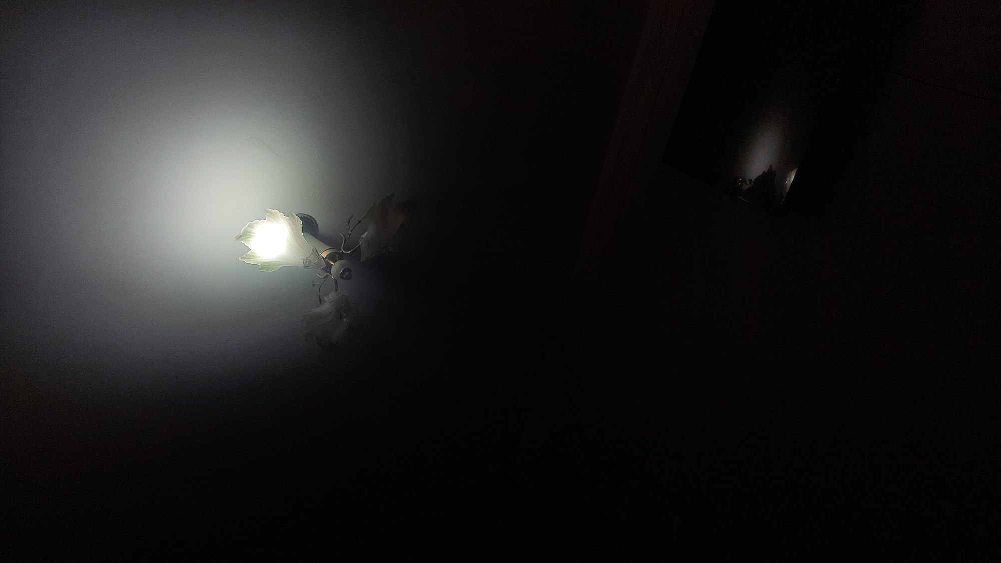 The image depicts a ceiling fan with a light fixture attached. The light is on and illuminates the area directly below it, creating a bright glow on the ceiling. The fan blades are visible, and the overall scene is dimly lit, with much of the image in shadow. There is a dark, reflective surface, possibly a piece of furniture or a floor, visible in the lower right portion of the image, catching some of the light. No people are present in the image. The setting is indoors, and no specific location in Bukhara, Uzbekistan, can be identified from the visual information provided.