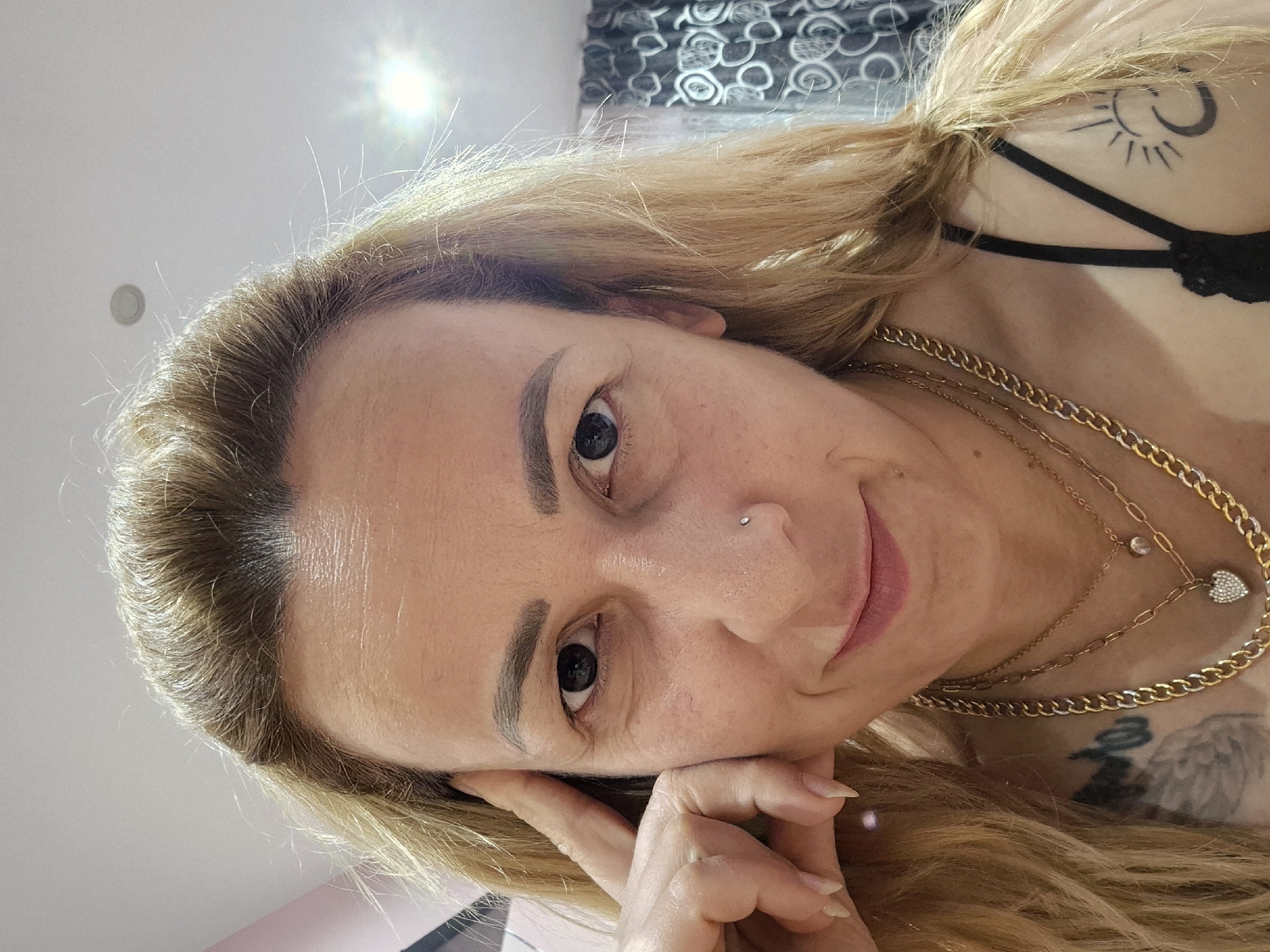 A close-up, angled portrait depicts a woman with blonde hair. She is resting her face on her hand. The woman is wearing gold necklaces. A tattoo is visible on her upper chest and another one on her shoulder. She has a piercing on the side of her nose. The location is unknown.