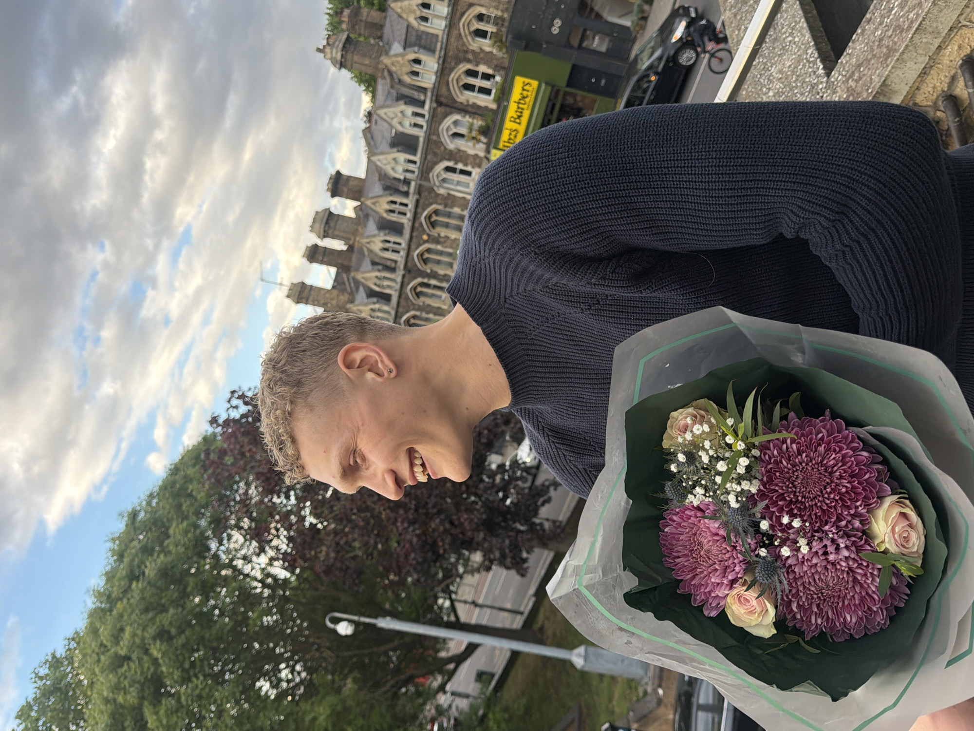 A rotated medium shot captures a smiling young Caucasian man with short, light-colored hair, wearing a dark blue knit sweater, holding a bouquet of flowers. The bouquet is wrapped in clear plastic over a dark green paper, containing purple chrysanthemums, light pink roses, and small white flowers. The background reveals a city scene: a multi-story brick building with arched windows and gabled roofs, a street with vehicles, a green sign reading "Ibz Barbers," trees, and street lighting. The cloudy sky indicates an outdoor setting. The location appears to be in London, United Kingdom, given the architectural style.