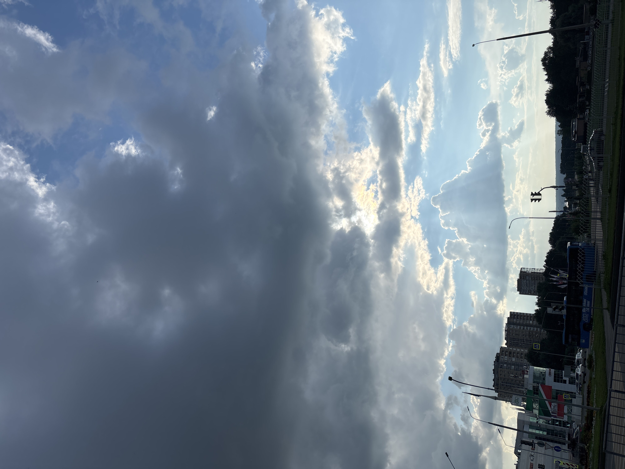 The image shows a cloudy sky over the city of Naberezhnye Chelny, Russia. The sky is a mix of dark storm clouds on the left and lighter, sunlit clouds on the right, with visible sunbeams breaking through. Below the sky, there are several modern buildings, including high-rise apartment buildings and what appear to be commercial structures with signage. A blue bus is visible on a road, along with cars. Trees line the horizon in the distance. The scene appears to be captured from a street-level perspective, looking towards the city skyline and the sky.