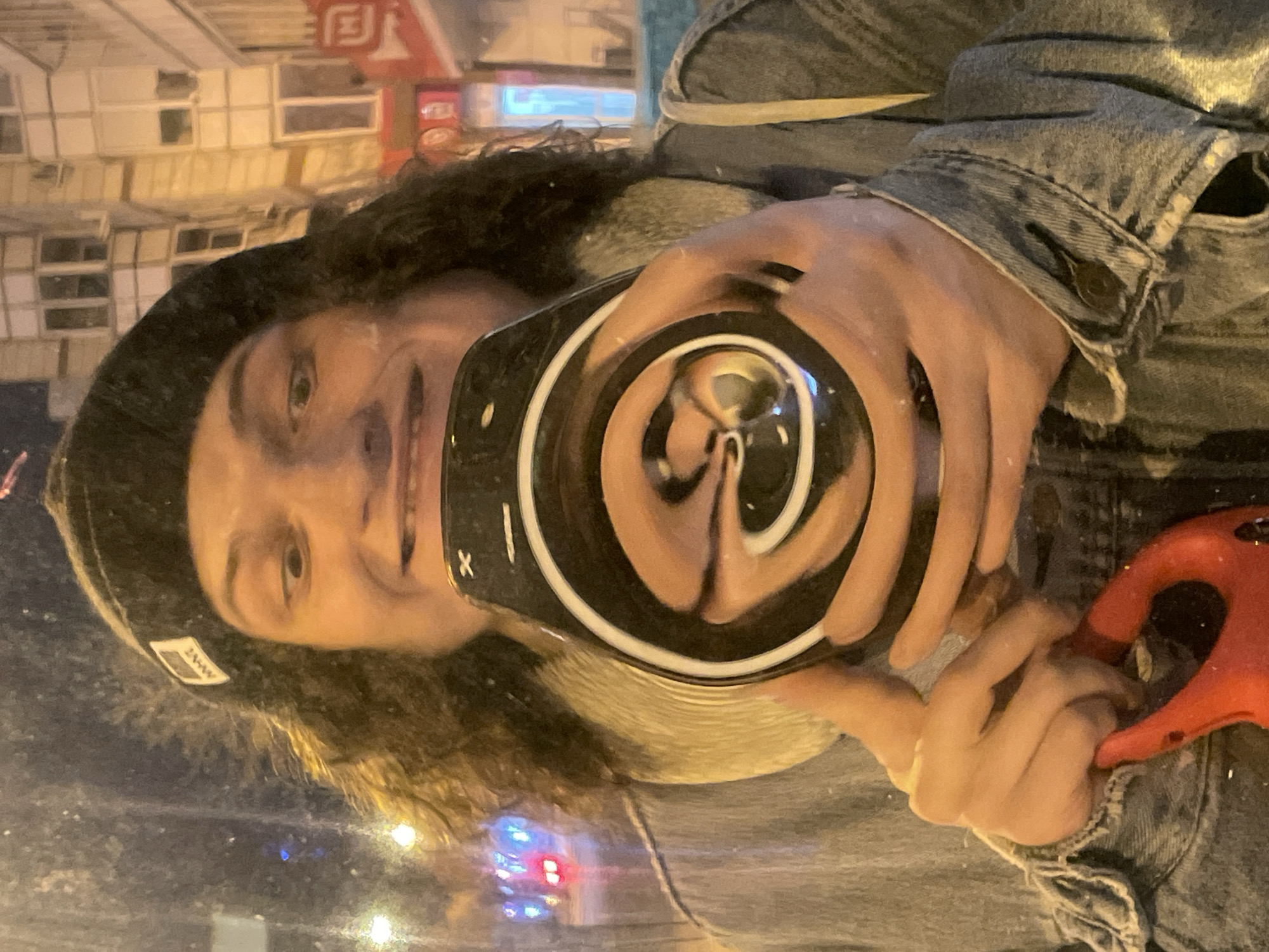 A person with curly hair and wearing a black beanie and denim jacket is taking a selfie in a distorted mirror, capturing their face and the reflection of the surrounding cityscape at night. The location is Yekaterinburg, Russia, indicated by a building with a distinctive red and white sign featuring Cyrillic script in the background. The person holds a red object in their other hand. The reflection in the mirror is swirling and heavily distorted. The city lights add to the ambience.