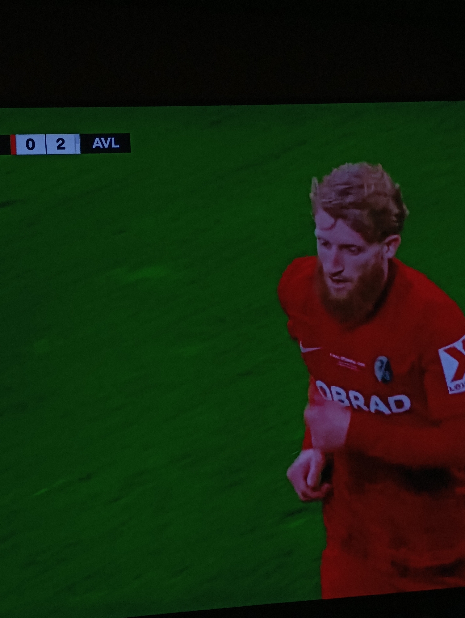 The image shows a soccer game in progress, with the score being 0-2, AVL leading. A red-haired, bearded soccer player is visible, wearing a red jersey with the word "OBRAD" on it. The game appears to be taking place on a green field. While it's impossible to determine the location specifically in Plovdiv, Bulgaria from this image alone, the player's jersey suggests it could be related to PFC Lokomotiv Plovdiv.