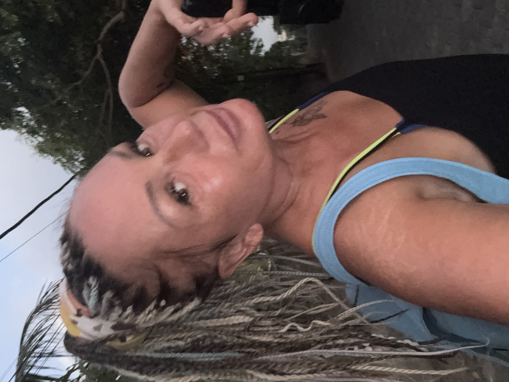 A medium shot, taken at dusk in Kuta, Indonesia, shows a woman from the chest up. She is facing the camera and looking towards it with her right hand behind her head. Her hair is pulled back into a high ponytail with dreadlocks and is adorned with a colorful bandana. She is wearing a black tank top with a blue and neon yellow strap. A blue backpack is visible around her left shoulder. The background shows trees and a dirt road.