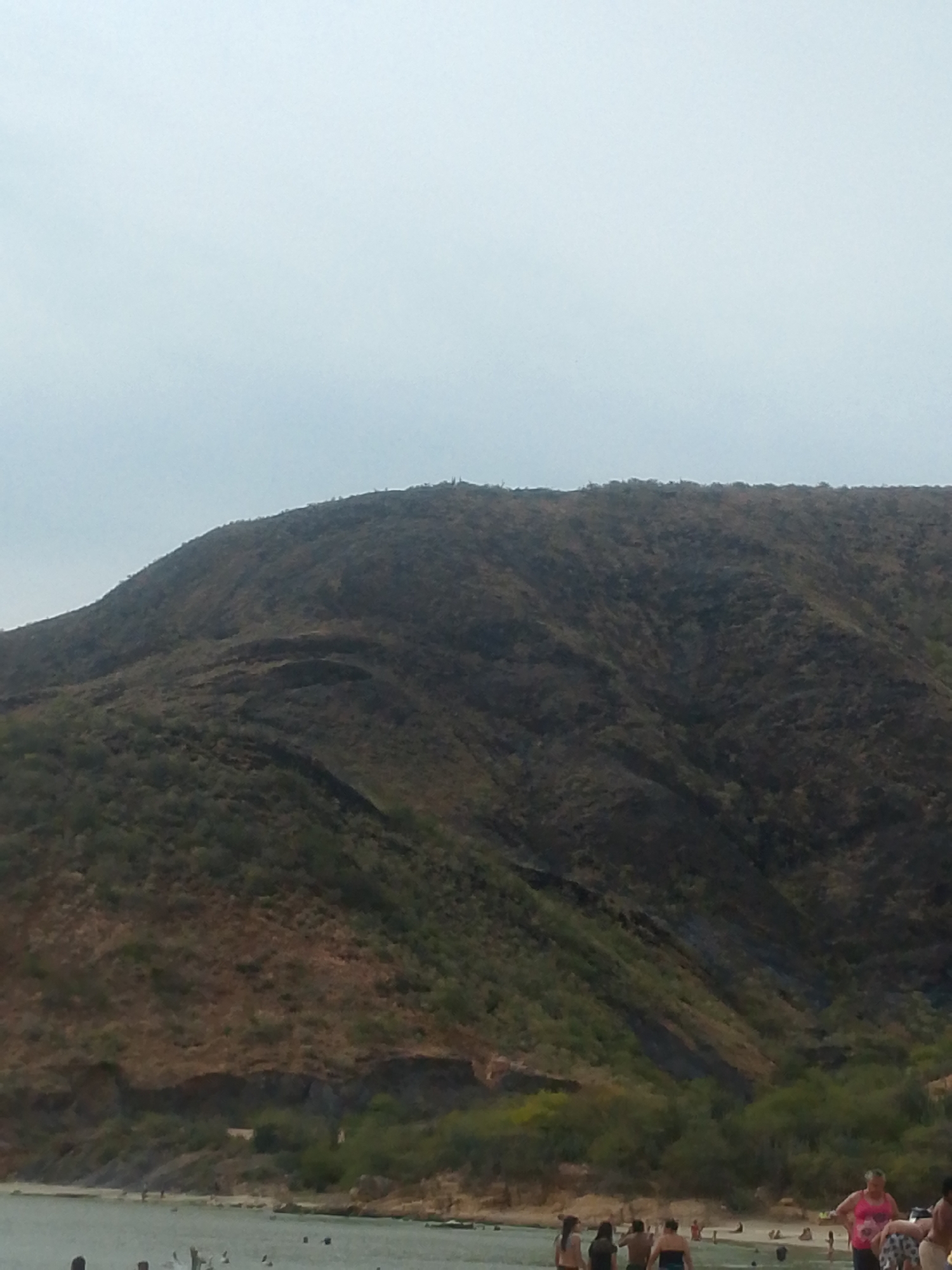 The image shows a beach scene in Juan Griego, Venezuela. A large, dry, sparsely vegetated hill dominates the background. The lower slopes of the hill transition into denser green vegetation. In the foreground, a beach area is visible, with many people either standing in the shallow water or walking along the shoreline. The sky is overcast and gray.