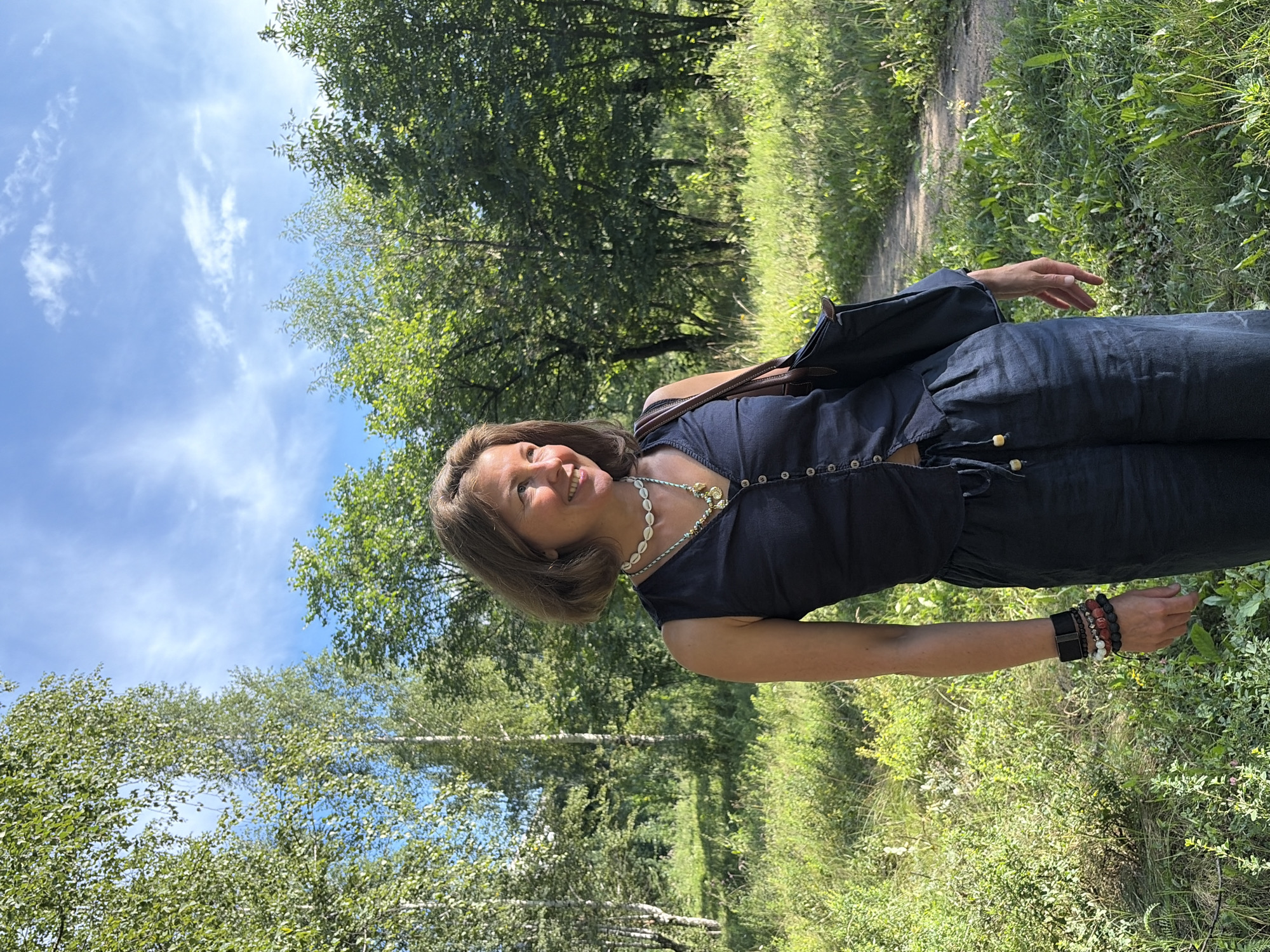 A woman stands in a grassy area with trees behind her. She is wearing a dark blue sleeveless shirt and dark blue pants. She has a necklace and bracelets. She is carrying a dark brown bag over her shoulder. The image was taken outdoors on a sunny day with a blue sky and some clouds. The location is unknown.