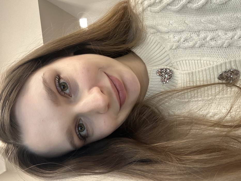 A fair-skinned woman with light brown hair looks towards the camera with a slight smile. She wears a cream-colored, cable-knit sweater adorned with silver heart-shaped buttons at the neckline. Her eyes are a distinctive shade of green. The image is slightly tilted, creating a casual and informal composition. The background is blurred and out of focus.