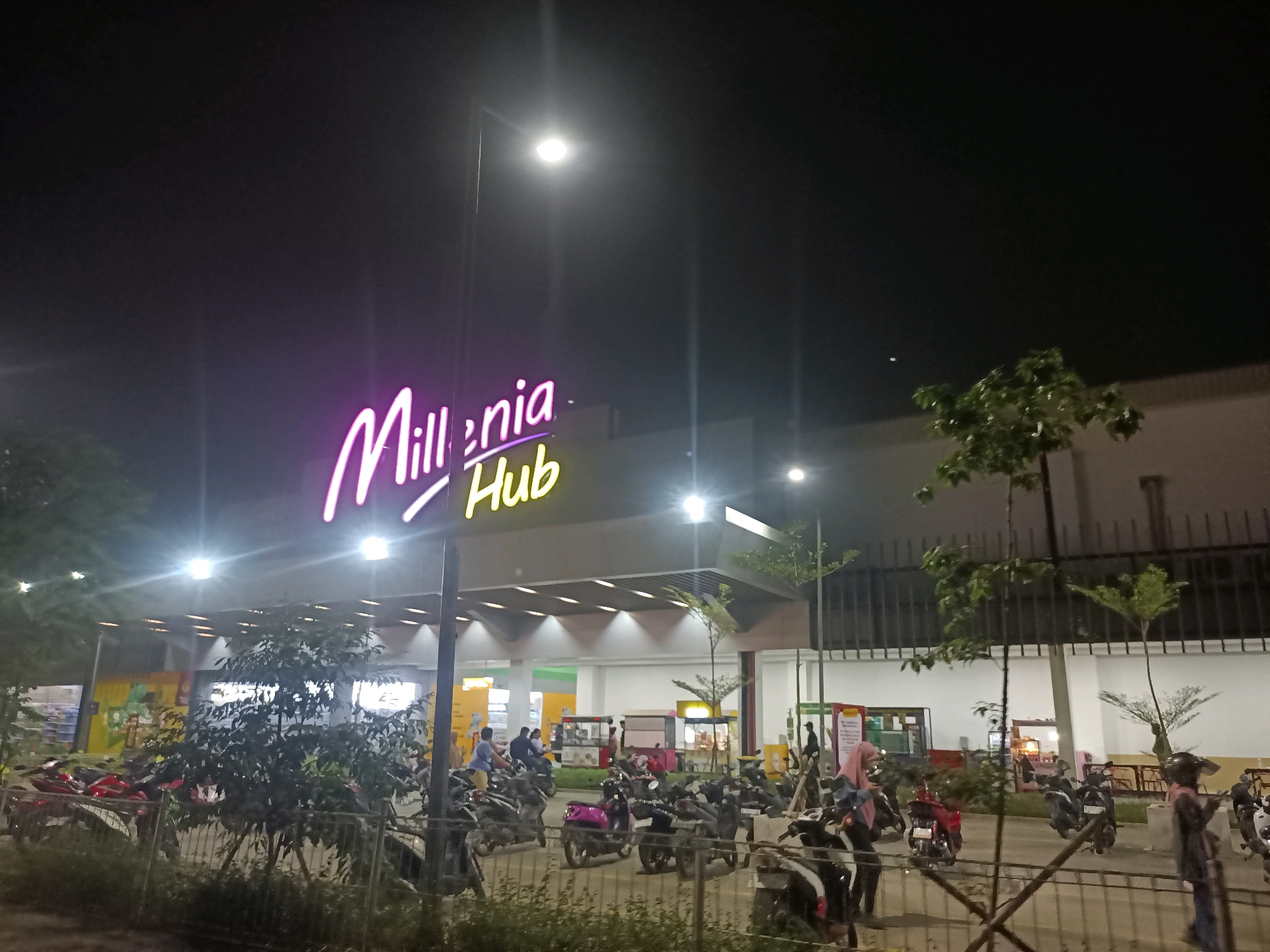 This is a night-time, eye-level, wide-angle photograph of the entrance to Millenia Hub, a commercial establishment in Telagamurni, Indonesia. The building's facade is illuminated by bright, modern lighting, and a neon sign spelling out "Millenia Hub" in purple and yellow is prominently displayed. In front of the building, a large number of parked motorcycles are visible, some with riders. People are present in the foreground and midground, some walking and others standing near the motorcycles. The area appears to be an outdoor parking lot or plaza in front of the commercial building. The overall atmosphere suggests a bustling commercial area at night.