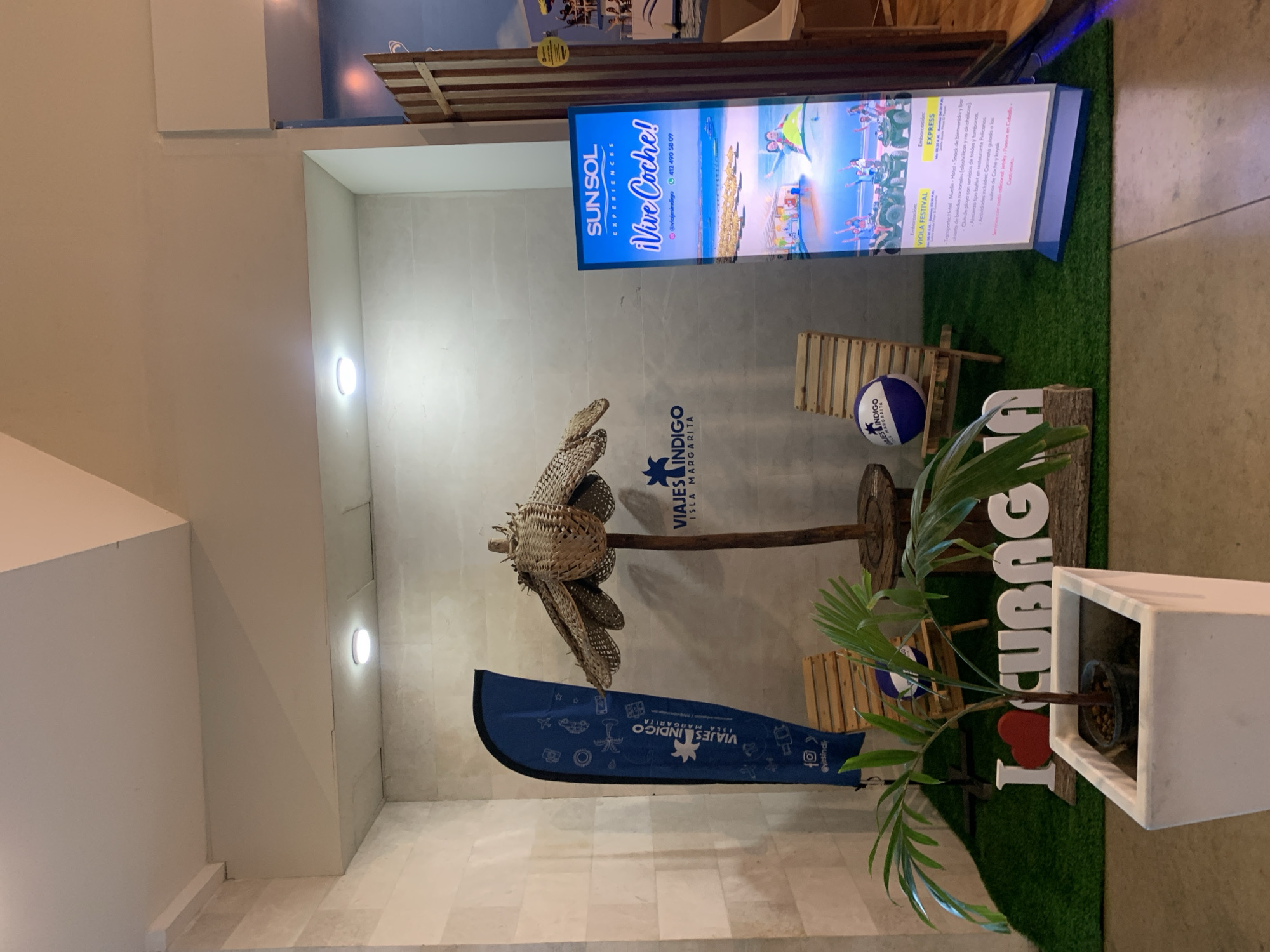 This media file captures an indoor promotional display, likely within a commercial space such as a travel agency or hotel in Pampatar, Venezuela. The scene features a themed setup designed to evoke a tropical vacation experience.
The central element is a bright, illuminated digital display advertising "SUNSOL EXPERIENCES" and encouraging visitors to "¡Vive Coche!" (Live Coche!), referring to Coche Island, near Isla Margarita. The screen shows vibrant images of people enjoying various water and outdoor activities. Detailed text on the screen outlines excursion packages, including "VIOLA FESTIVAL" and "EXPRESS," which offer transport, hotel stays, welcome snacks and open bar (alcoholic and non-alcoholic), beach club services (tents and sunbeds), buffet lunch at Pelicanos restaurant, and activities such as guided walks to the Coche salt flats, kayaking, horseback riding, and quad bike tours. Contact information, "@viajesindigo" and a phone number (412 490 58 09), are also visible.
The ground of the display is covered with artificial green turf, mimicking grass. Decorative elements include a large, intricately woven, light-brown, flower-like sculpture mounted on a wooden pole. Below it, a small wooden beach chair sits next to a blue and white beach ball. A small green plant, possibly a palm frond, is potted in a white rectangular planter. Behind the plant, partially obscured, are large white letters forming "I ❤️ CUBAG," likely part of "I love Cubagua," another island near Margarita. A blue pennant flag or banner, bearing the logo and text "VIAJES INDIGO ISLA MARGARITA" along with various travel-related icons, stands on the left.
The background consists of light grey walls, with spotlights in the ceiling and a circular wall sconce providing illumination. The overall ambiance is inviting and highlights recreational opportunities in the Venezuelan Caribbean. No people are visible in the image.