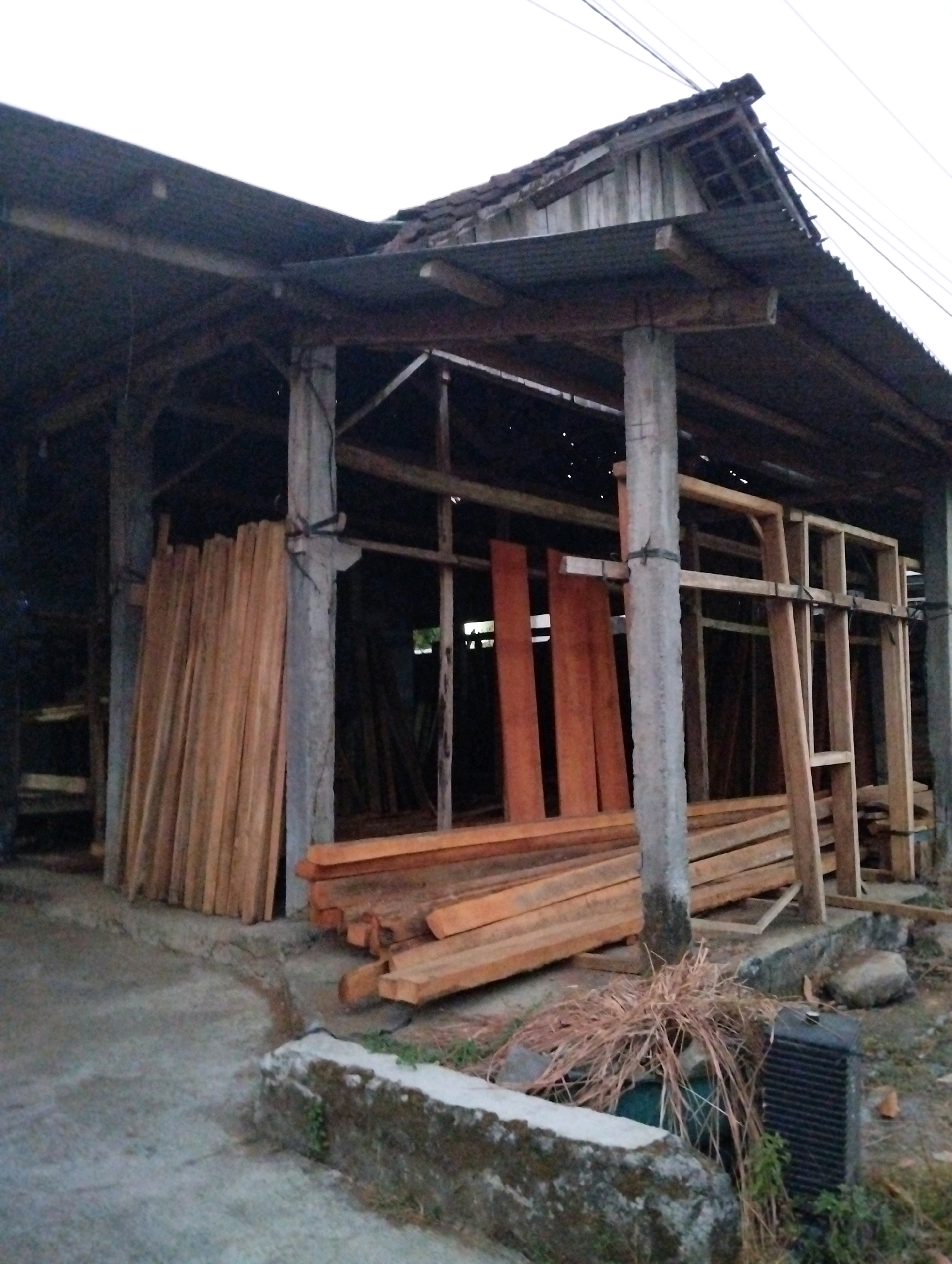 An outdoor, low-angle, medium shot depicts a woodworking facility in Salatiga, Indonesia. The facility, a covered open-air structure with a corrugated metal roof and concrete pillars, houses a large collection of lumber. The lumber is stacked in various orientations, some upright against a pillar, others in neat piles on the concrete ground. The wood appears to be raw and unfinished, varying in color from light tan to reddish-brown. Behind the lumber, more unfinished wood, possibly for window frames, is visible, secured to a wooden support structure. A pile of dried grass and a black, rectangular object, possibly a fan, are situated to the right of the lumber piles. The ground is a rough concrete surface. The overall scene suggests a functional, outdoor workshop environment.