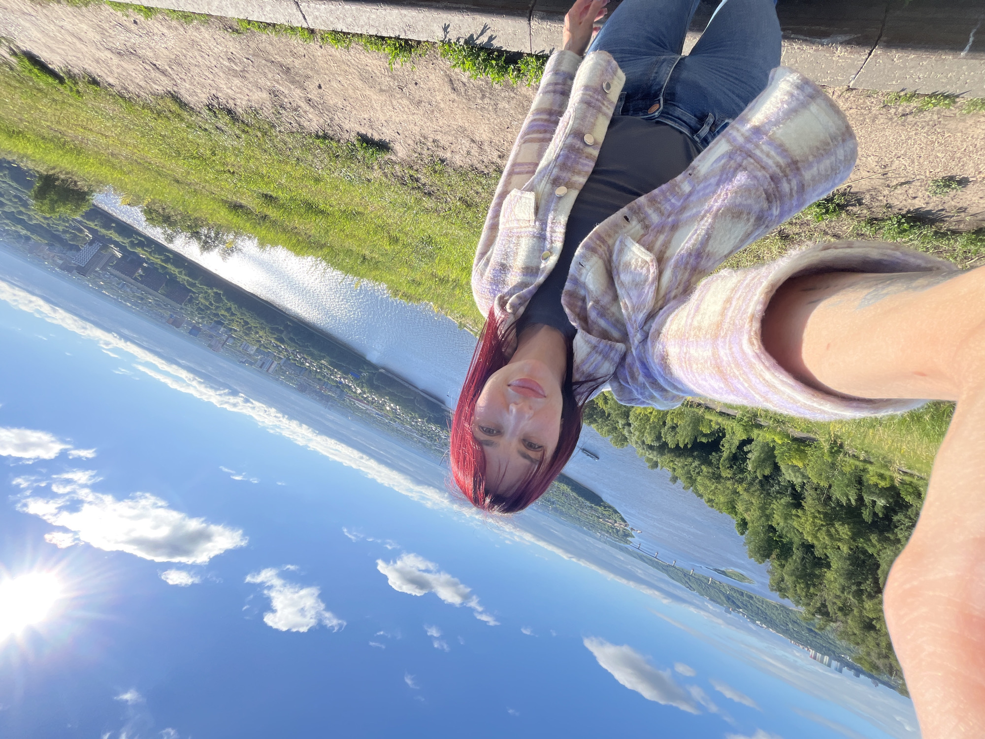 In Nizhny Novgorod, Russia, on a sunny day with a clear blue sky dotted with white clouds, a woman with vibrant red hair takes a selfie on a grassy hillside overlooking a wide river and a city. She is wearing a plaid shirt in shades of white, purple, and pink, a dark t-shirt, and blue jeans. The sun shines brightly in the upper right corner of the frame. The background shows the expanse of the river, with a bridge and a ship visible in the distance, and a cityscape with buildings nestled among trees. The woman is positioned in the foreground, her arm holding the camera out to capture the selfie and the scenic view.