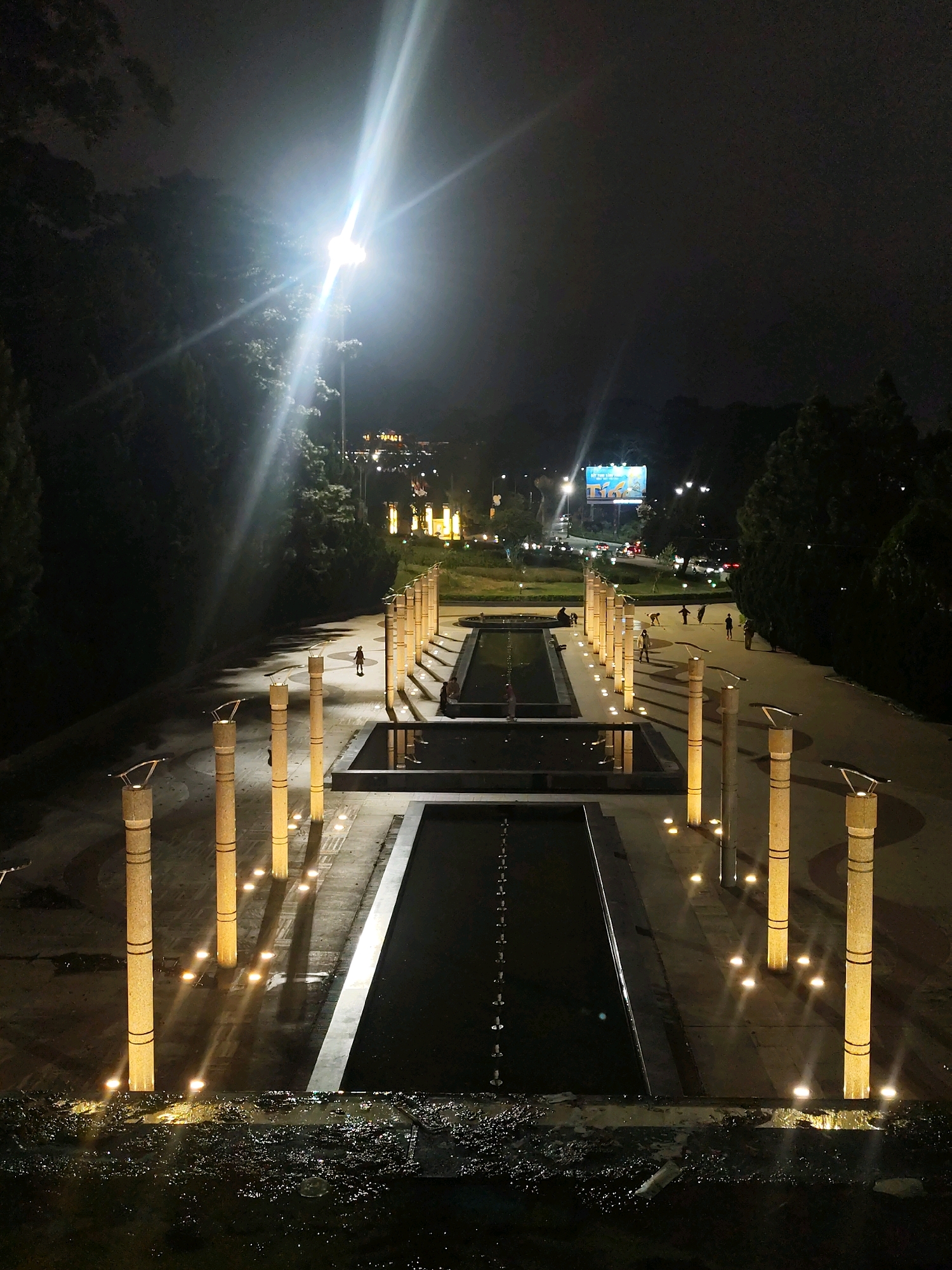 The image depicts a nighttime scene at the Da Lat Flower Garden in Da Lat, Vietnam. The garden features a symmetrical layout with long, rectangular water features, likely fountains or reflecting pools, running parallel to a paved walkway. Tall, cylindrical pillars, illuminated by lights at their bases, line the walkway. The path is also dotted with small, ground-level lights that cast pools of illumination.
In the background, the city of Da Lat is visible with illuminated buildings and streetlights. A large billboard displaying advertising is also present. The overall atmosphere is serene and illuminated by artificial lights against the dark night sky. Several individuals are scattered throughout the scene, some walking along the path, others appearing to be resting near the water features or on benches. The foreground shows a dark, wet surface with debris, suggesting a potentially water-logged area or a close-up view of a fountain edge. The central focus is the illuminated walkway and water features, leading the eye towards the distant city lights.