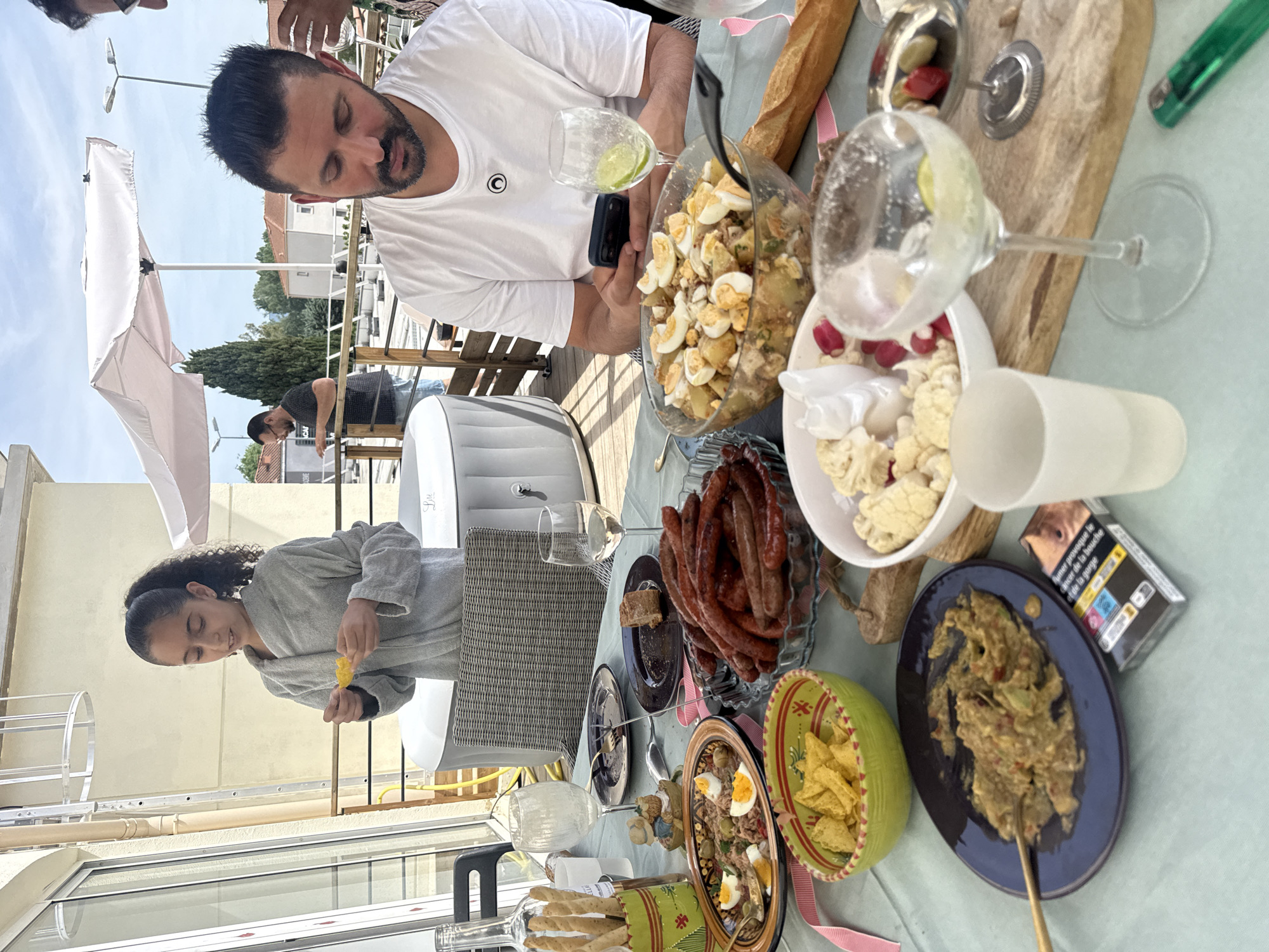 An outdoor social gathering is captured on a sunlit patio or terrace. The scene is centered around a table laden with various dishes, suggesting a meal or a casual party.
In the foreground, a man with dark hair and a beard, wearing a white t-shirt, is seated at the table, looking down intently at his smartphone. Beside him, a large glass bowl holds a potato salad topped with hard-boiled egg halves. A young girl with dark, curly hair tied back, dressed in a grey robe, is visible further down the table, holding a piece of yellow food.
The table is covered with a light blue tablecloth and features a spread of food: a bowl of cauliflower florets with radishes, a plate piled with cooked sausages (possibly merguez), a baguette, a bowl of tortilla chips, and a salad dish containing tuna, olives, and hard-boiled eggs. Another plate holds a chunky dip, likely guacamole. Glasses, some with drinks like a lime-garnished one, and bottles (one holding breadsticks) are also present.
The background shows a bright, clear sky and an open white umbrella providing shade. Further back, residential buildings with tiled roofs and green trees are visible. An inflatable hot tub and a ladder can also be seen in the outdoor setting. The overall atmosphere appears relaxed and casual, under bright daylight.
Visible text on a cigarette packet on the table reads "FUMER PROVOQUE LE CANCER DE LA BOUCHE ET DE LA GORGE" (Smoking causes mouth and throat cancer).