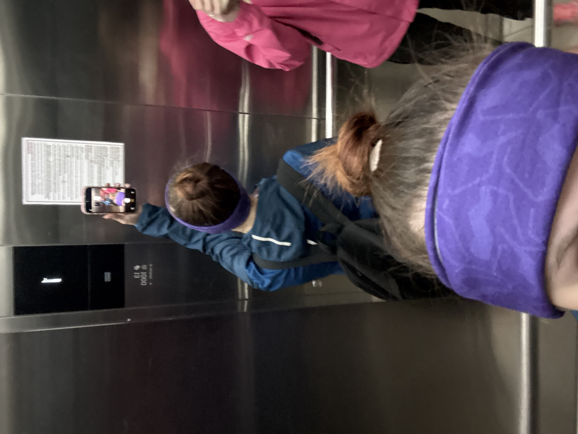Inside an elevator, a person in a blue jacket and purple headband is taking a picture with their phone of the elevator's safety information. A second person with a purple headband and another wearing a pink jacket are also present. The elevator's panel indicates it is on the first floor and has a weight capacity of 1000 kg. The location is likely Perm, Russia, based on the 13+ weight capacity indication (which is common to Russian elevator plates).