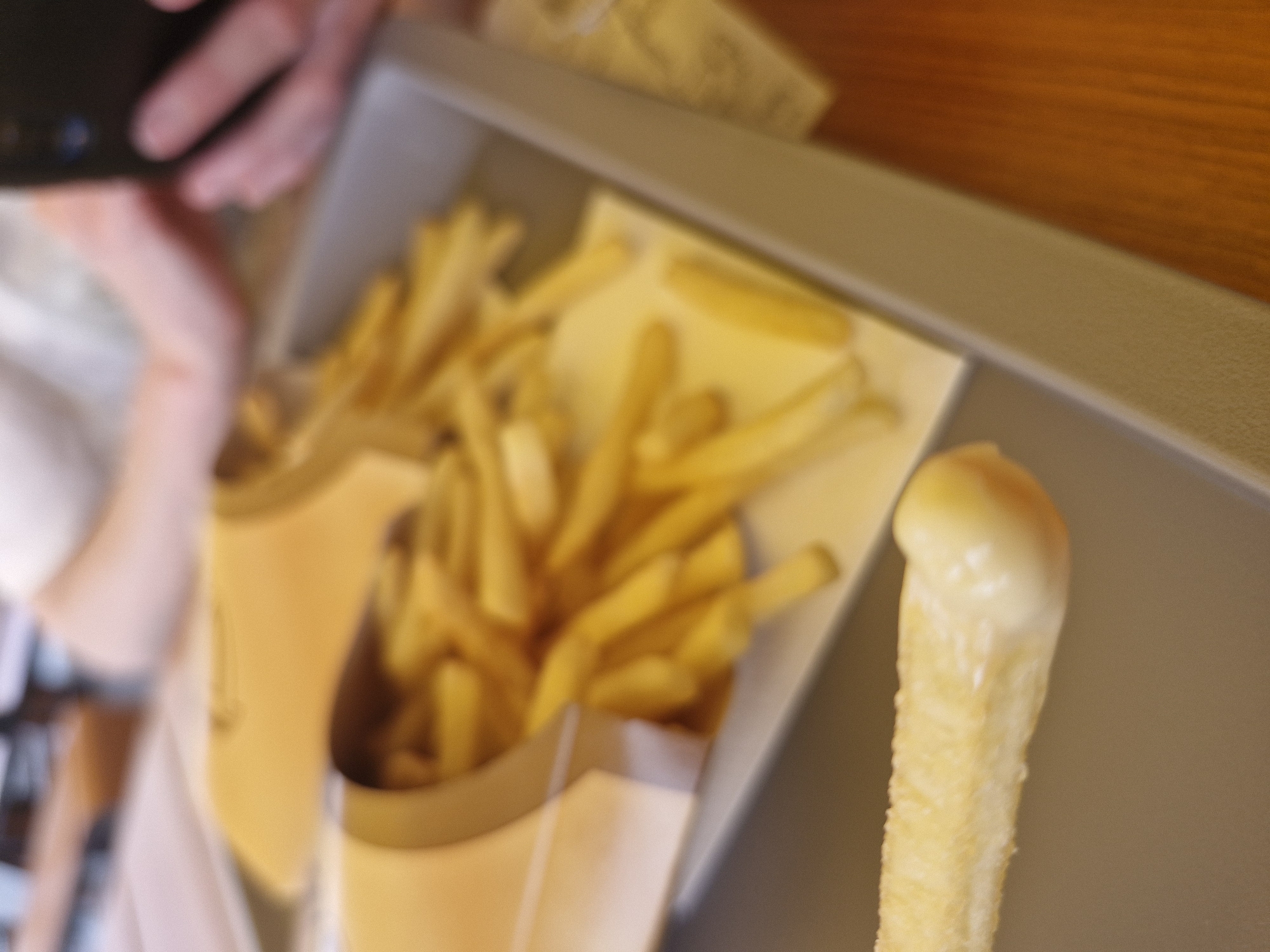 An overhead shot shows a serving tray filled with french fries and one fry dipping sauce. The tray is placed on a wooden table. Two hands are visible in the background, one holding a mobile phone. The scene is indoors, likely in a restaurant.