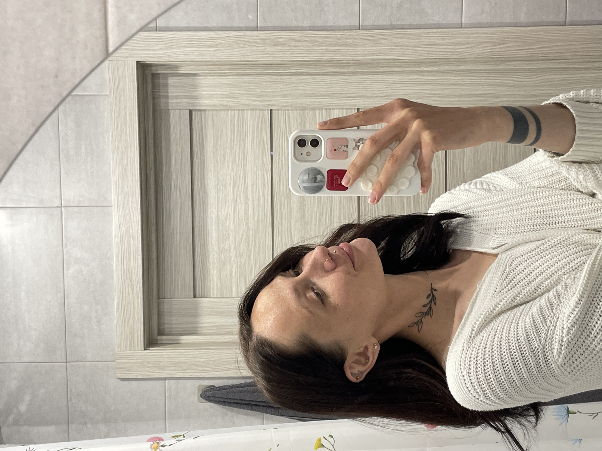A woman is taking a selfie in a bathroom. She is holding a white phone with a colorful case. She is wearing a white sweater. She has dark hair and visible tattoos on her wrist and neck. The bathroom has light-colored tiled walls and a light wood-framed mirror. There is a patterned shower curtain at the bottom of the frame. The location is not identifiable as Izhevsk, Russia, based on the visual information.