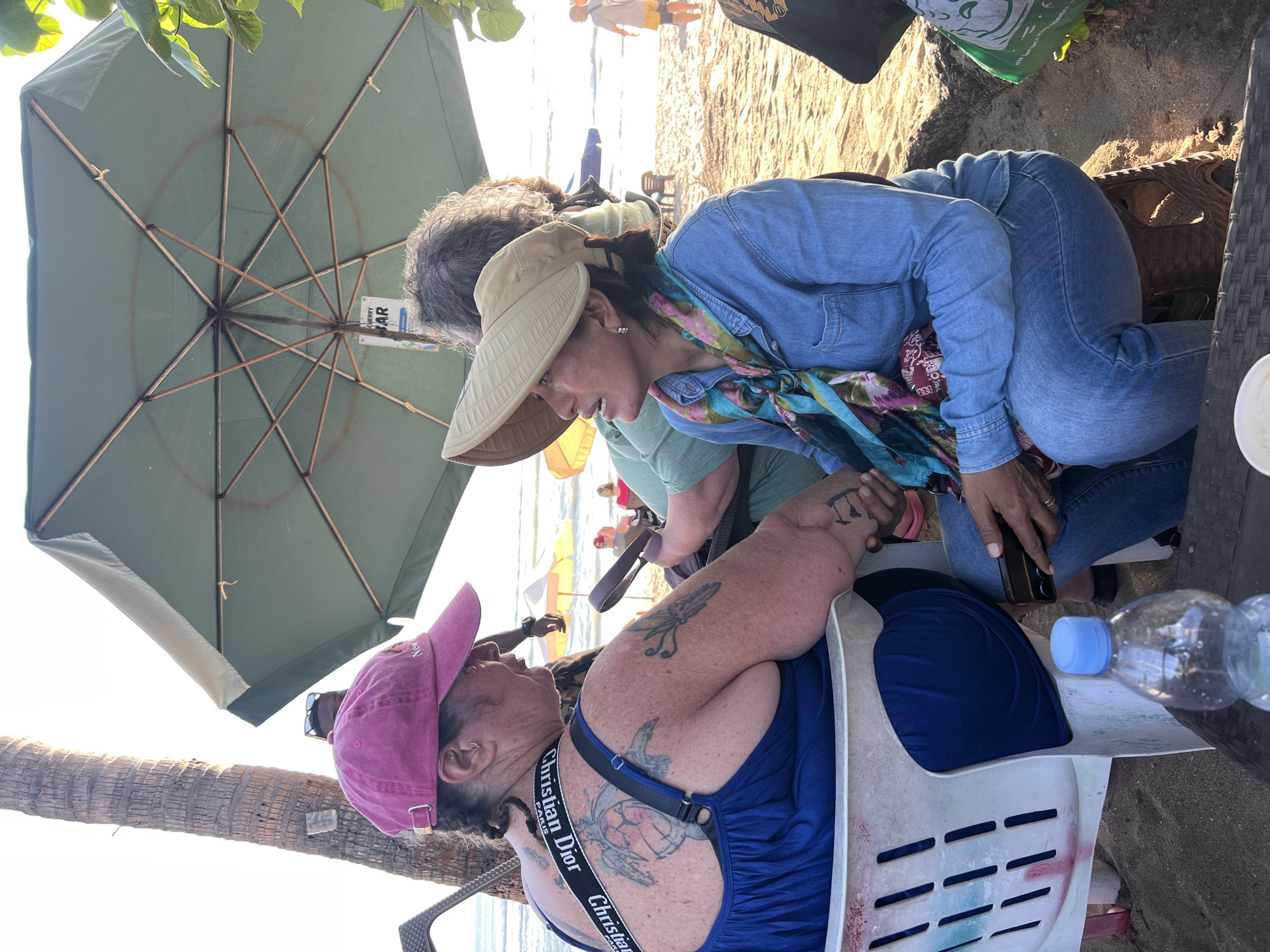 Two women are sitting at tables on a sandy beach in Kuta, Indonesia, under a large green umbrella. The woman on the left is wearing a pink baseball cap and a blue dress. She has extensive tattoos on her back and arms, including a turtle design. The woman on the right is wearing a tan sun hat and a denim shirt with a colorful scarf. She is looking to her left and appears to be in conversation with the other woman. In the foreground, a plastic water bottle is on a table. The background shows the ocean, other beachgoers, and palm trees. The lighting suggests it is late afternoon or early evening.