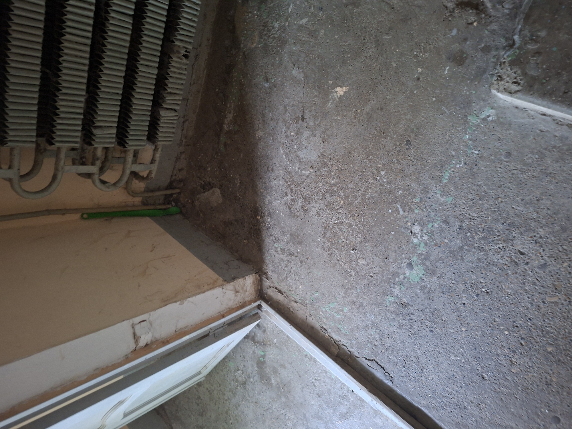 The image depicts a close-up, eye-level view of a dimly lit, unfinished interior space. In the foreground, a rough concrete floor is visible, showing cracks and scattered green paint splatters. To the left, a partially opened white door with ornate detailing is visible, its frame appearing damaged and peeling. Behind the door, a concrete wall continues. The right side of the image is dominated by a large, grey, corrugated metal structure, likely a radiator or heat exchanger, mounted against a cream-colored wall. A thin green object, possibly a broom handle or a piece of pipe, leans against the wall near the radiator. The environment appears utilitarian and possibly in a state of disrepair, suggestive of a basement, utility room, or an older building. The location in Kazan, Russia cannot be determined from the visual information provided.