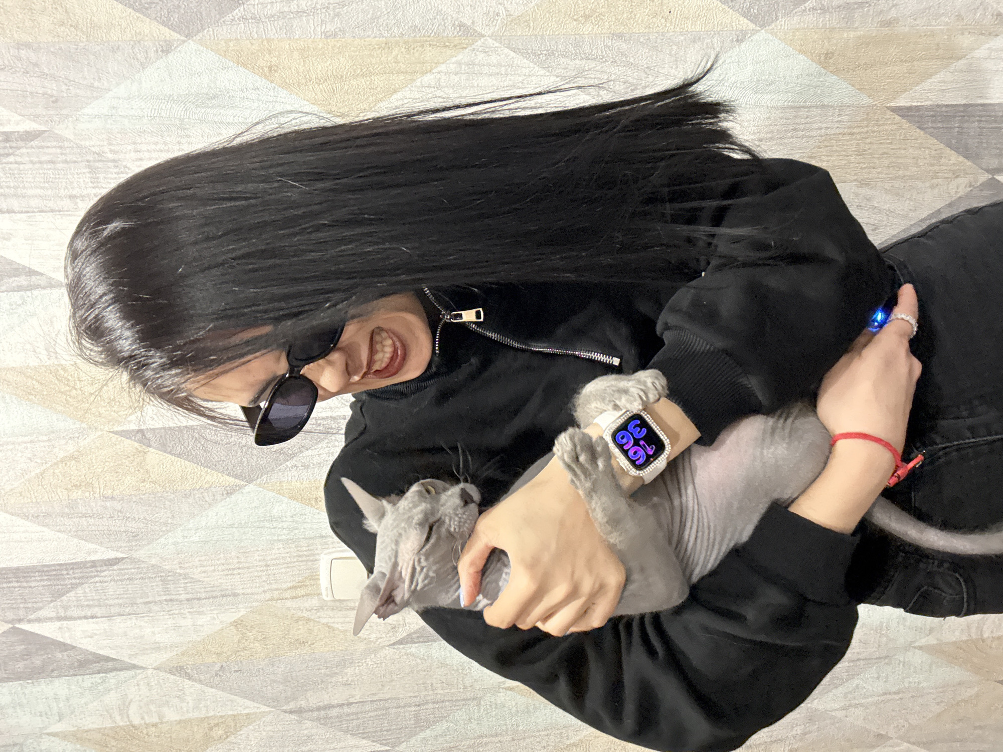 A woman with long, straight, black hair, wearing sunglasses and a black bomber jacket, is holding a gray hairless cat. She is smiling with her teeth showing. On her left wrist, she has a smartwatch with a white bedazzled band displaying the time as 3:36. On her right wrist, there's a red string bracelet. The background is a geometrically patterned wall. The location is unknown; Votkinsk, Russia cannot be confirmed.