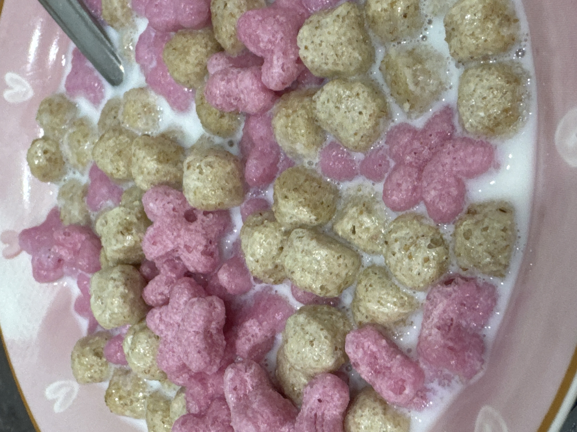 This is a close-up, overhead shot of a bowl of cereal. The cereal consists of two types of pieces: small, round, light brown pieces and larger, pink, flower-shaped pieces. Both types of cereal are submerged in milk, which has a slightly foamy texture on the surface. The bowl itself appears to be pink and has a subtle heart pattern visible on its surface. A silver spoon is partially visible at the top left of the frame, suggesting that someone is either about to eat or is in the process of eating the cereal. The lighting is bright, indicating it is likely daytime, and the setting appears to be indoors. There is no visible text in the image. The overall impression is one of a simple, pleasant meal.
