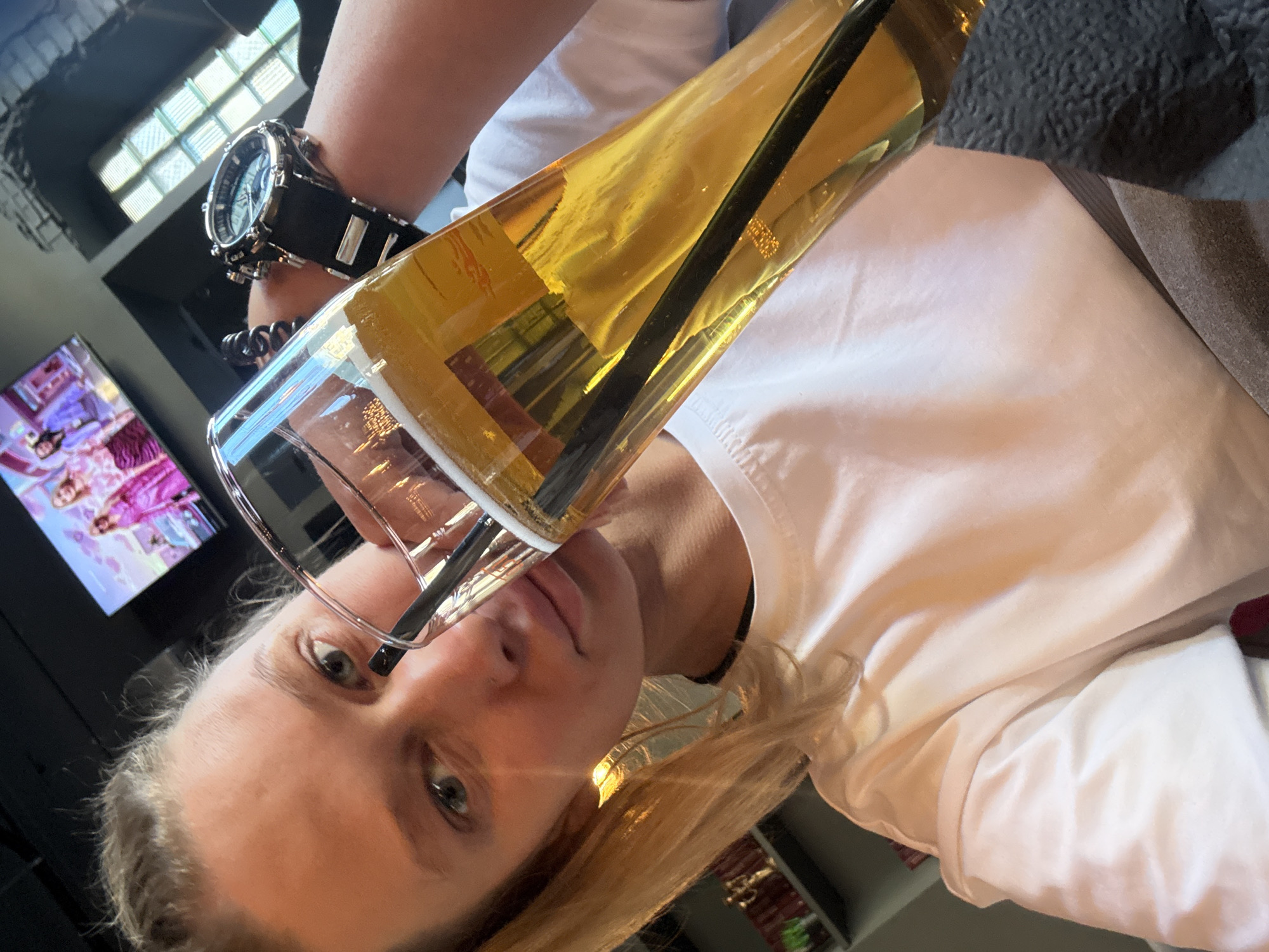 A blond woman is holding a large glass of amber liquid, likely beer, with a black straw, close to her face. She is wearing a white t-shirt and a large black watch. A television is visible in the background, as well as the city of Perm, Russia.
