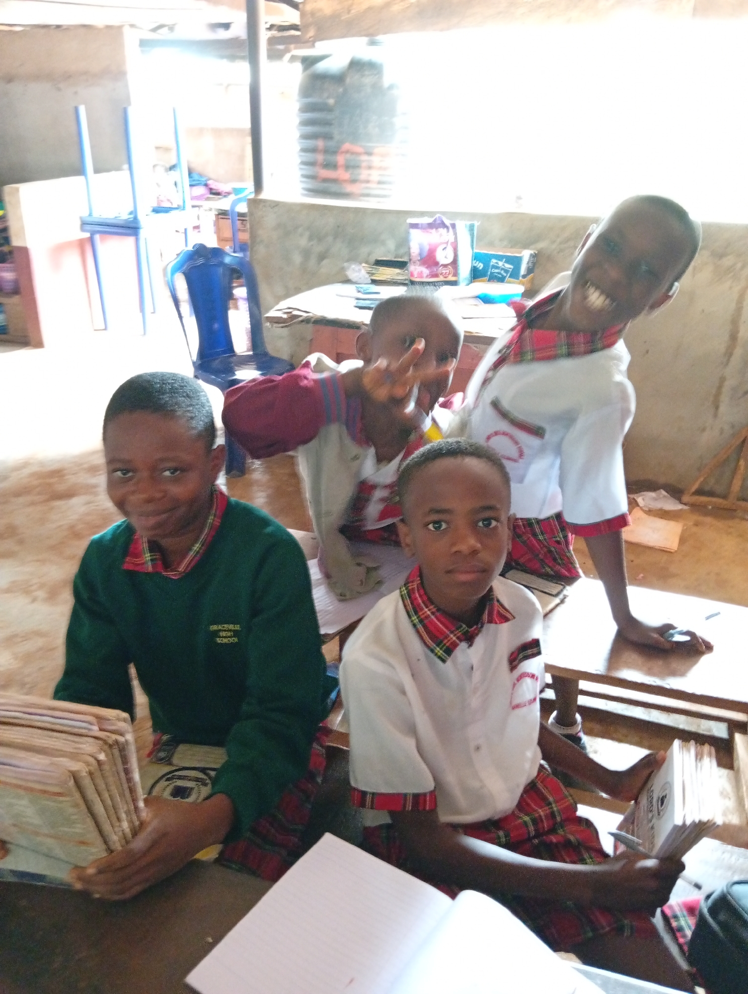 The image shows four young children indoors. One child wears a dark green sweater with "Gracaville High School" visible on the left breast. Another child is in a white shirt with red plaid trim on the collar and sleeves and red plaid pants. A third child wears a white shirt with red plaid trim, and the fourth child wears a red sweater with white sleeves and red plaid trim. The children are gathered around a table. One child is holding a stack of books. The setting appears to be a simple, perhaps rural, classroom. The location is Unknown, Unknown.