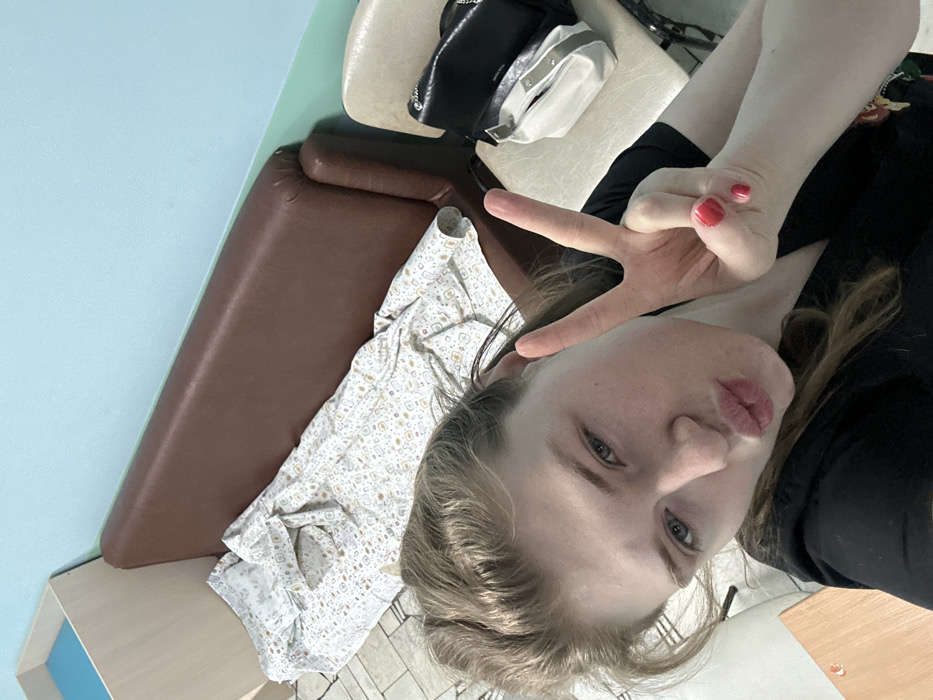 A woman with light hair and blue eyes takes a selfie, pouting her lips and holding up a peace sign. She wears a black top. Behind her is a light blue wall, a brown padded bench, and a white dress with a subtle pattern. There are also two purses on a beige surface. The image is rotated counter-clockwise.
