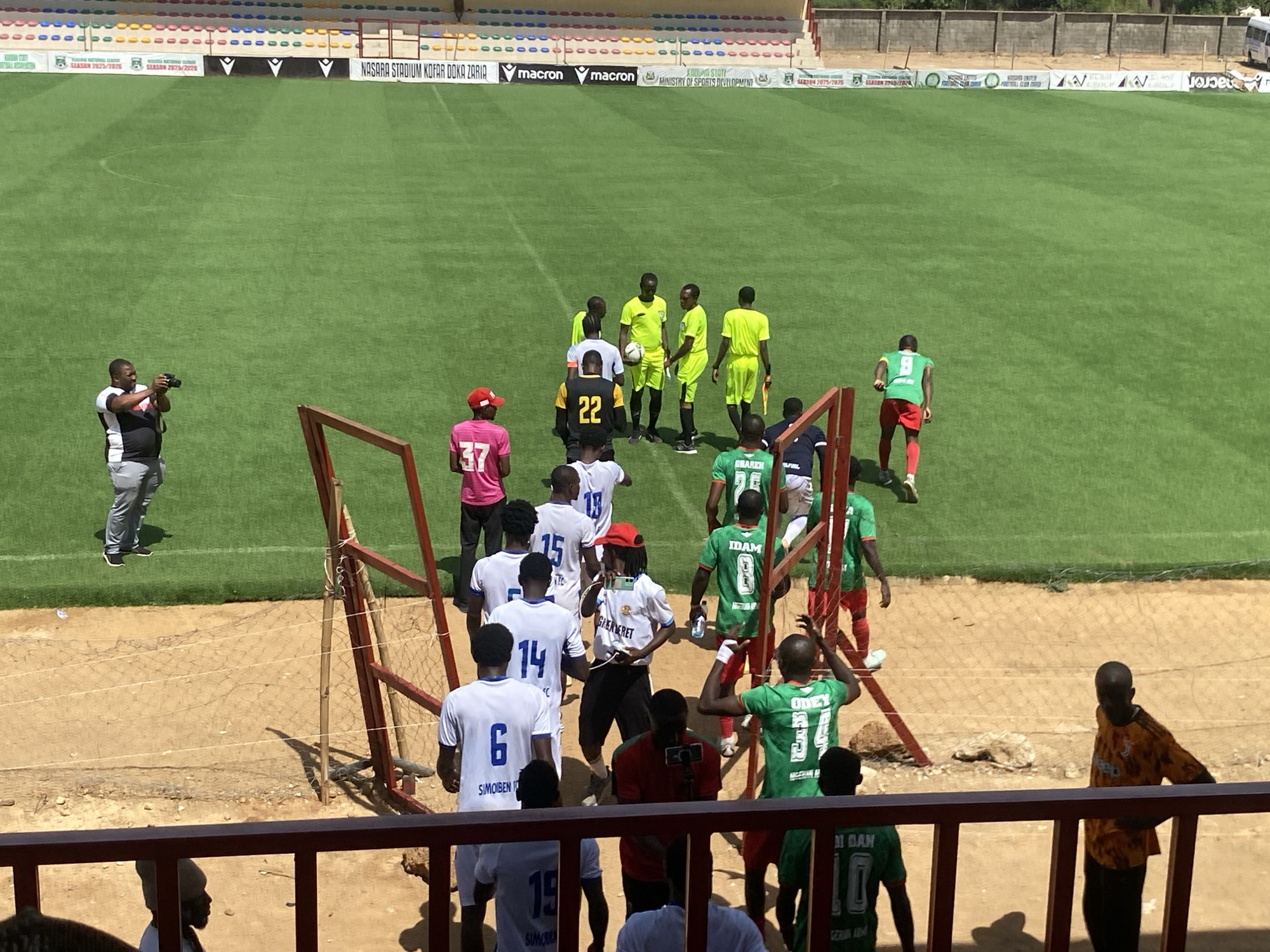 The image depicts a soccer match at Nasara Stadium Kofar Doka Zaria in Tudun Wada, Nigeria. Soccer players wearing various jerseys (white with numbers 6, 14, 15, 19; green with numbers 8, 10, 26, 34; black with number 22; pink with number 37; orange) are entering the field through a metal-framed opening. A man is taking a picture of the scene. Referee in a yellow jersey. The field is green, and in the background, there are stadium seats and advertising banners for "macron" and "Ministry of Sports Development."