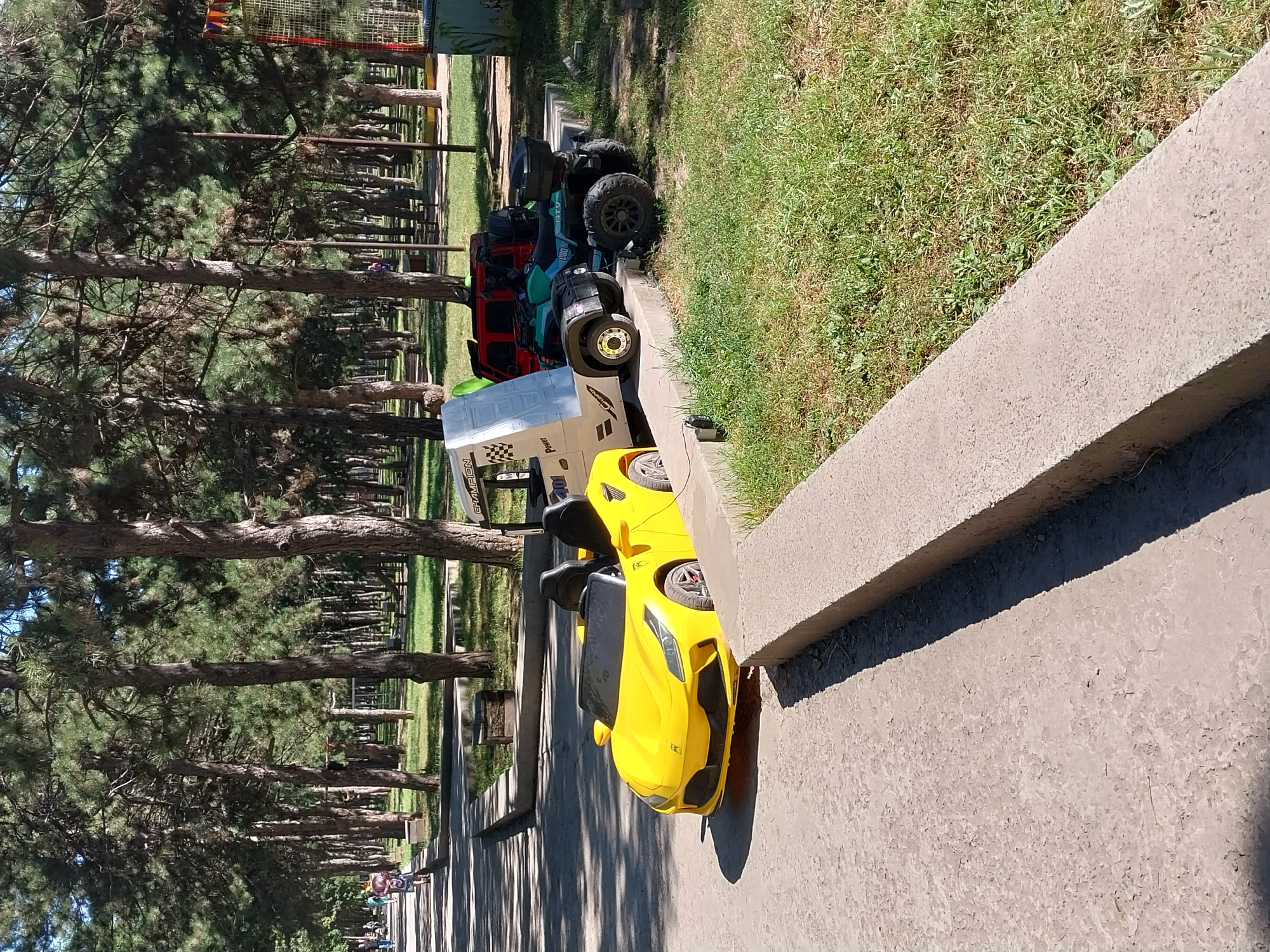 The image shows a park in Dnipro, Ukraine. In the foreground, a yellow toy sports car is parked on a concrete surface. Behind it, a toy truck, a green all-terrain vehicle (ATV), and a red toy SUV are also parked on a raised concrete platform. The vehicles are positioned in front of a line of trees, suggesting they are part of a play area or rental service within the park. In the background, there are more trees, grassy areas, and a playground structure. Several people, including children, are visible in the distance, walking along a path. The overall scene depicts a sunny day in a recreational outdoor space.