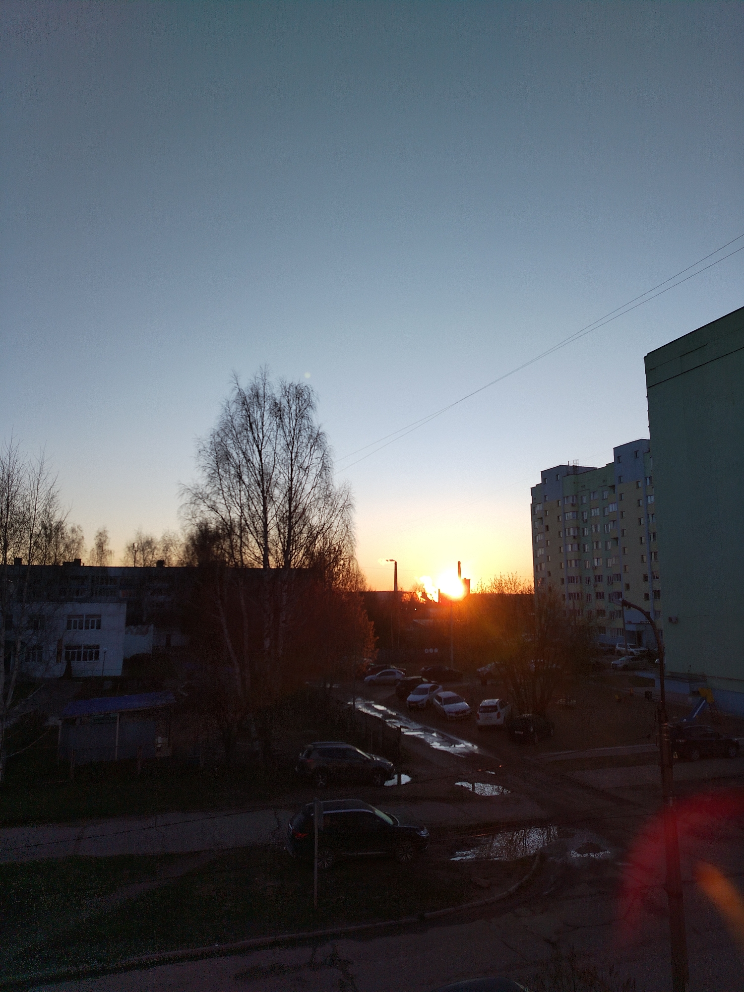 This image depicts an urban or suburban residential landscape at either sunrise or sunset. The sun, a bright orange orb, is low on the horizon, partially obscured by buildings and trees on the left side, casting a warm glow across the lower sky. The upper sky is a clear, light blue.
In the foreground and midground, multiple apartment buildings are visible. A large, light green/teal building occupies the right side of the frame, and a multi-colored apartment block (featuring blue, yellow, and green sections) is situated further back and to the left. Numerous bare-branched deciduous trees, including a prominent birch-like tree in the center, are silhouetted against the sky, some catching the warm light of the sun.
The lower half of the image shows a paved area, likely a parking lot or residential courtyard, where several cars are parked. Multiple puddles are scattered across the dark ground, reflecting the ambient light and indicating recent rain. Thin power lines stretch across the upper part of the sky. No people are visible, and no specific activity is taking place, conveying a sense of quiet and tranquility. There is no visible text in the image.