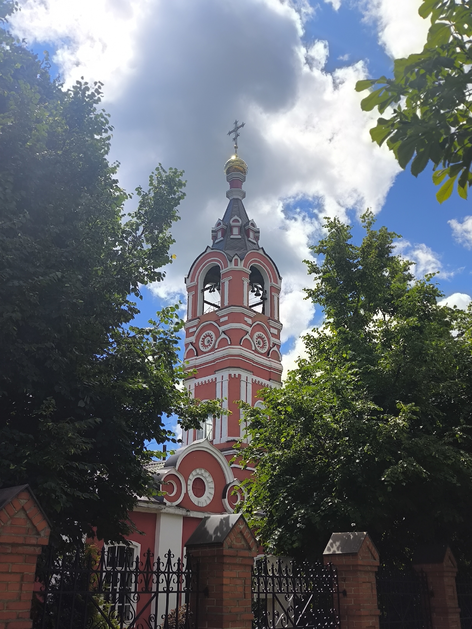 A bell tower of a red and white church is visible through lush green trees against a partly cloudy blue sky. The bell tower has a golden dome topped with a cross and features arched openings with bells. The church building has a red facade with white decorative elements. In the foreground, a black metal fence with intricate scrollwork is supported by brick pillars. The scene is located in Taldom, Russia.