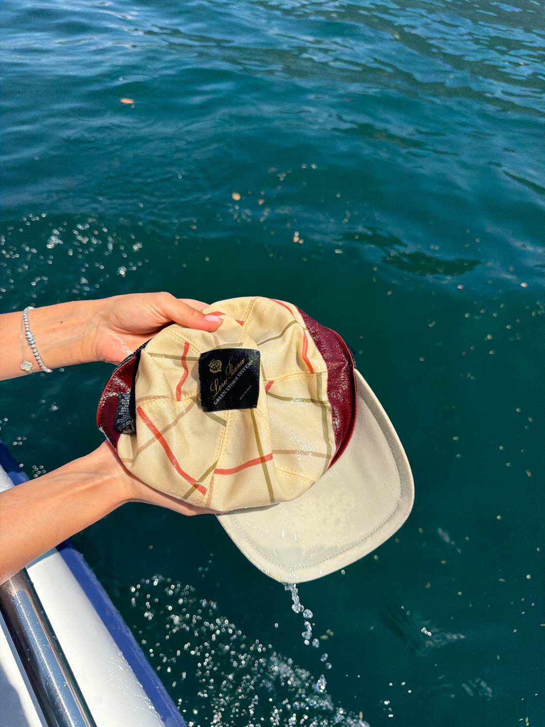 In the city of Nesso, Italy, a person is holding a beige and maroon plaid baseball cap over dark blue water. The cap has a beige brim and a beige interior with a tag that reads "Loro Piana" and "Green Storm System." Water is splashing from the brim of the cap into the lake below. The person's hands and arms are visible, with one hand wearing a silver bracelet. The background is dominated by the deep blue water, with some small debris floating on the surface. The scene appears to be taking place on a boat, as a white railing is visible in the lower left corner.