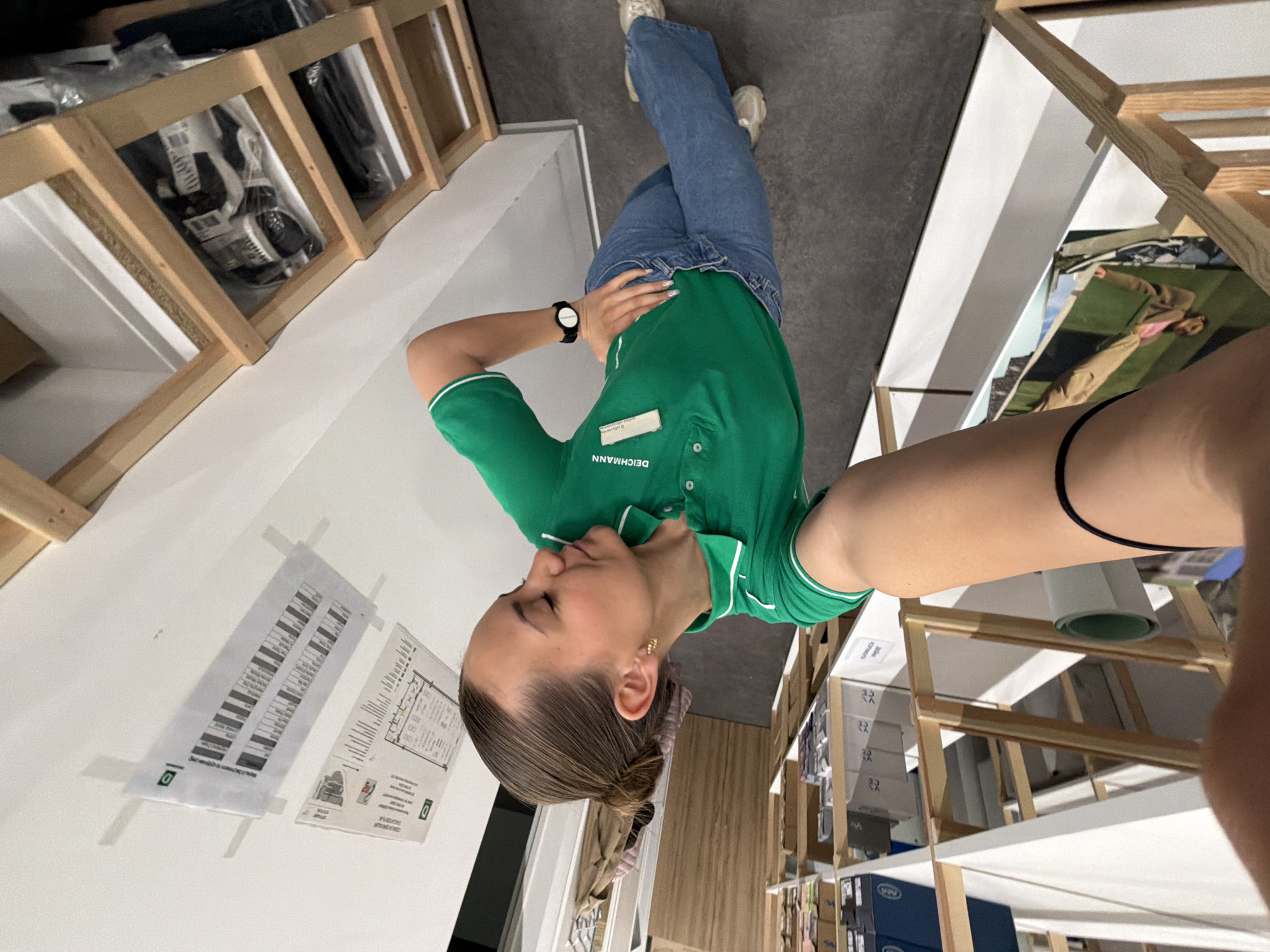 A young woman in a green DEICHMANN employee polo shirt takes a selfie in what appears to be a DEICHMANN shoe store. She is wearing blue jeans and beige sneakers. Her hair is tied back. She is wearing a black wristwatch on her left wrist and a gold earring. Shelves of shoe boxes are visible behind her on both sides. Evacuation plans are attached to the wall. The location is possibly in Sofia, Bulgaria, given the presence of DEICHMANN, a retail chain with stores in Bulgaria, and the building layout suggested by the evacuation plans.
