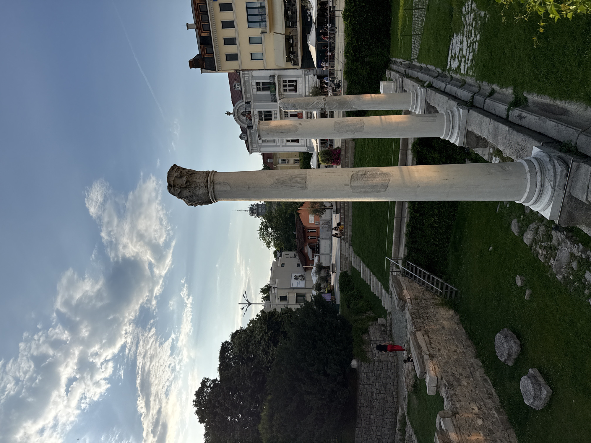 The image depicts an outdoor archaeological site in the city of Plovdiv, Bulgaria, during the late afternoon or early evening. The scene is illuminated by warm, directional sunlight, casting long shadows.
In the foreground and midground, several tall, weathered white marble columns with visible carvings, including one with a Corinthian capital, stand vertically on stone bases. These columns are part of ancient ruins, bordered by low, rough-hewn stone walls. A rectangular grassy area is present between these walls and the columns, with scattered large rocks. To the left, a person in a red top and dark pants walks on a cobblestone path adjacent to a stone wall, with a small child nearby. A wooden ladder extends downwards into the excavated area.
Towards the center-right, stone steps ascend a grassy embankment. Beyond these steps and the ruins, a paved area with outdoor cafes and restaurants is visible, populated by several individuals seated under white umbrellas. Modern, multi-story buildings with balconies and varied architectural styles, including light yellow and grey facades, form the background. Signs, including "caffetteria," are visible on these buildings.
On the left side of the image, dense green foliage and large trees obscure parts of the background, though a multi-armed streetlight is visible above the trees. Further in the distance, a tall, spired structure or tower is discernible on a hill. The sky overhead is light blue with scattered white and grey clouds; a faint contrail is present in the upper right quadrant.