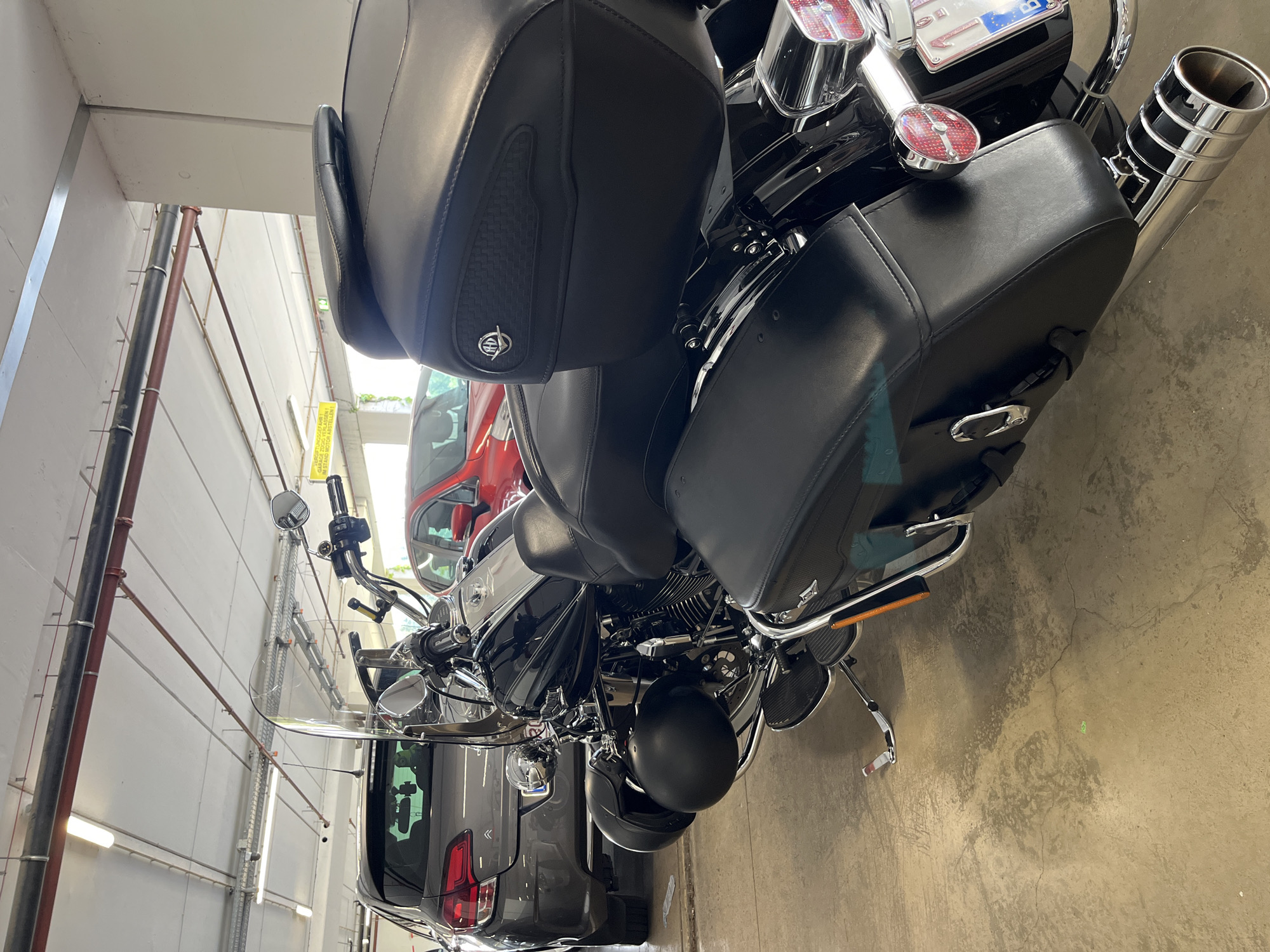 In a covered parking garage, a black Harley-Davidson motorcycle is parked facing away from the camera. The motorcycle has leather saddlebags and a large backrest. A grey car is parked behind and to the left of the motorcycle, and a red car is parked behind and to the right. The garage has a concrete floor and ceiling with exposed pipes and fluorescent lighting. A yellow sign with German text is visible on the ceiling. A license plate with "B" is visible on the back of the motorcycle. The image was taken in Weiswampach, Luxembourg.