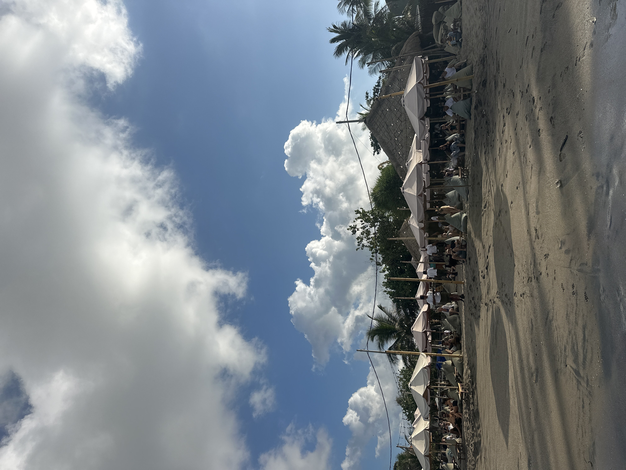 This image depicts a vibrant beach scene at a resort in North Kuta, Indonesia. In the foreground, the sandy beach shows footprints and the edge of the ocean. The middle ground is filled with rows of cream-colored umbrellas providing shade for beachgoers. People are seated on beanbag chairs and lounge chairs, appearing relaxed and enjoying the environment. Some staff members are visible, possibly serving guests. Beyond the seating area, there are thatched-roof structures, indicative of the resort's architecture, and lush palm trees. The background is dominated by a bright blue sky with scattered fluffy white clouds, suggesting pleasant weather. String lights are visible above the umbrellas, hinting at evening ambiance.