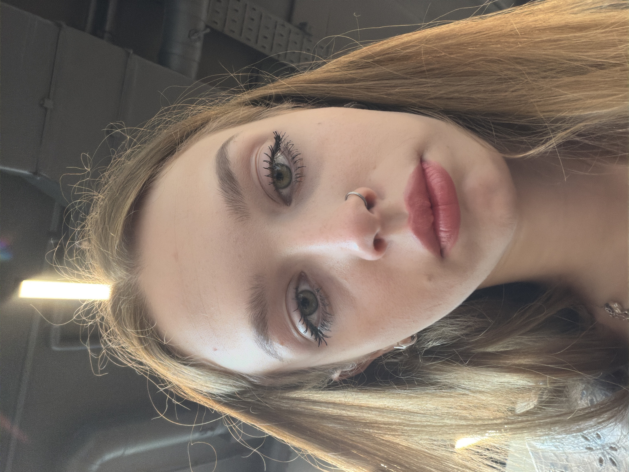 A close-up, selfie-style portrait captures a young woman with long, blonde hair. She has green eyes, is wearing mascara, and has a silver hoop nose ring. Her lips are painted a muted pink. She is wearing a silver necklace. The background is out of focus and appears to be an indoor environment with overhead lighting and some industrial-looking ceiling fixtures.