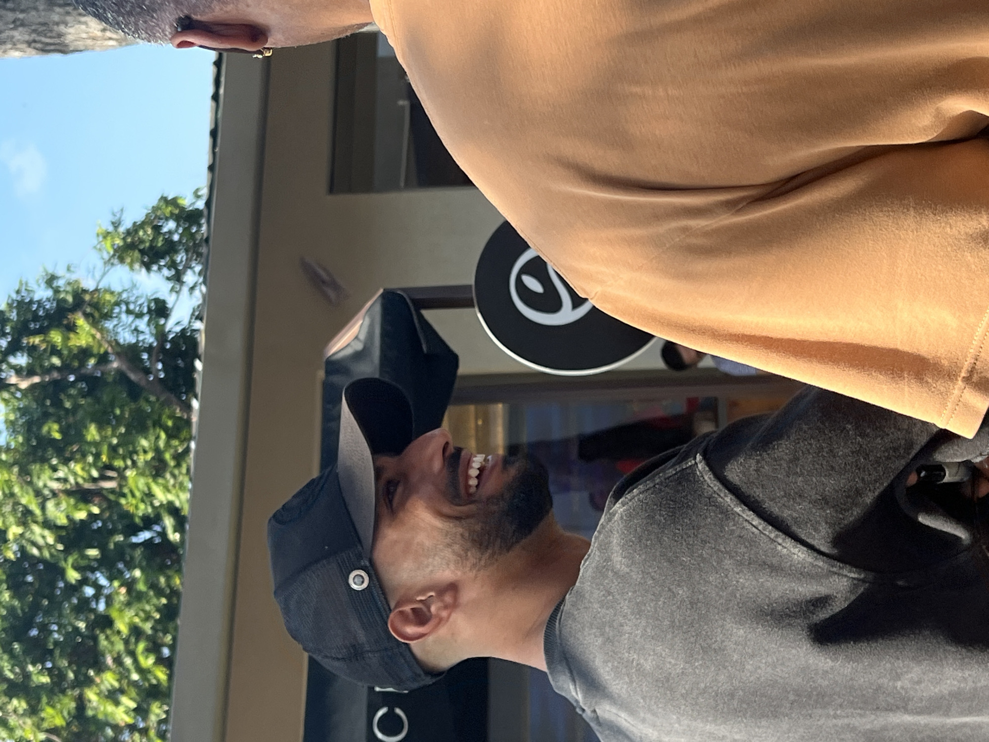 The image shows two men in an outdoor urban setting, possibly in Mengwi, Indonesia. The man on the left, wearing a black baseball cap and a grey t-shirt, is smiling and looking to his right. He has a beard and is in profile view. The man on the right, wearing an orange t-shirt, is seen from the back, with only his shoulder and the back of his head visible. A round black sign with a white graphic is visible between the two men. The background shows trees and the facade of a building. The lighting suggests it is daytime.