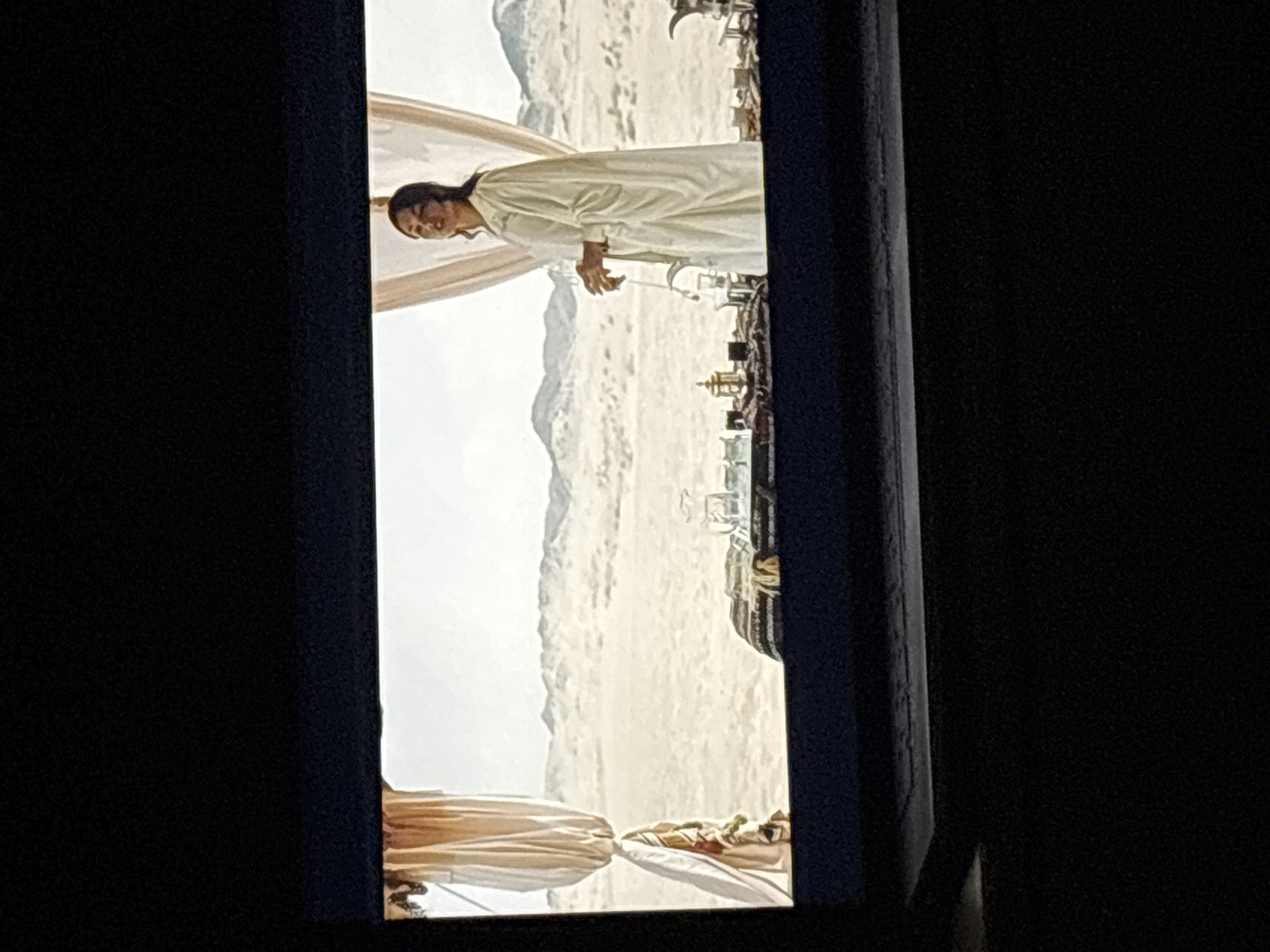 The image shows a screen displaying a scene with a woman in a long, cream-colored robe standing in front of a table set with several small glasses and what appears to be a decanter. Behind her, draped curtains frame a vast desert landscape with rolling sand dunes and distant mountains under a hazy sky. The woman has dark hair and is looking to her right, with her hands clasped in front of her. The table is covered with a patterned cloth, and various small containers are arranged on it. The scene is viewed from a slightly elevated angle, suggesting it's being watched on a display device, possibly a laptop, given the dark border and the lower portion of the image which appears to be the device's frame. The context of Budva, Montenegro is not visually present in the image.