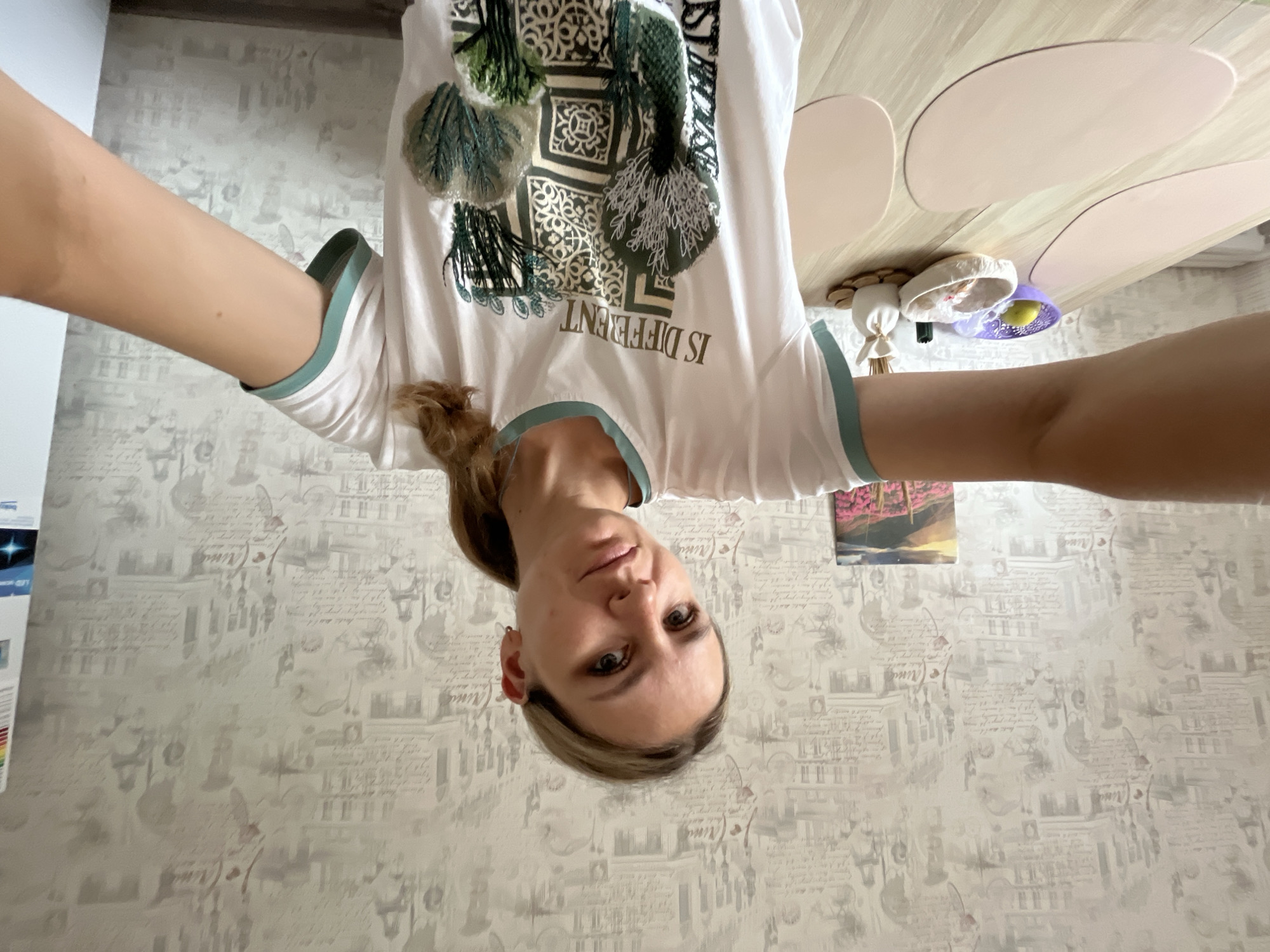 A young woman with blonde hair pulled back in a ponytail, wearing a white t-shirt with a green trim, takes a selfie in a room with patterned wallpaper. She is standing in front of a wooden table with several pink oval placemats. On the table, there is a purple bowl containing a lemon, a white woven basket with some items inside, and a small white vase with dried reeds. In the background, a framed picture of a landscape with pink flowers and mountains is visible on the wall. The scene appears to be an indoor domestic setting, potentially a kitchen or dining area. The location is specified as Pionersky, Russia.