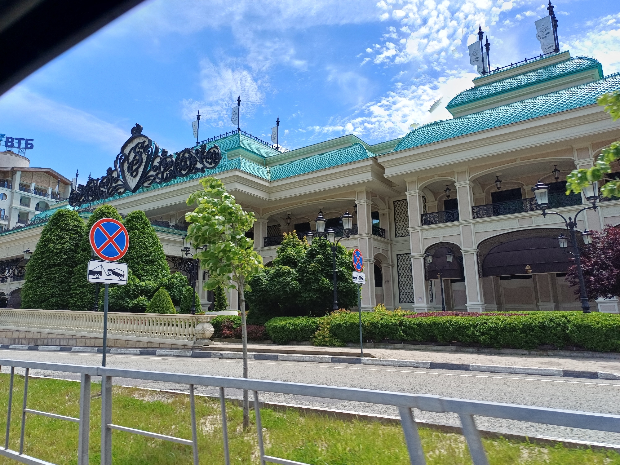 The image captures a grand, ornate building with a distinctive turquoise tiled roof, likely a casino or luxury resort, viewed from across a road in Estosadok, Russia. The building features cream-colored facades, intricate black wrought iron decorations, and multiple arched entrances or windows adorned with dark awnings and black lanterns. Above the main entrance, a large, elaborate crest in black and white adds to its opulent appearance.
In the foreground, a gray metal railing separates the viewer from a road with asphalt paving and a curb marked with white and gray stripes. Along the sidewalk leading to the building, a neatly trimmed hedge and various bushes, including some with reddish foliage, line the property. A young tree with light green leaves stands prominently near the road, and another larger, dense green tree is visible behind it.
Two street signs are clearly visible: a circular red sign with a blue cross indicating "no stopping or standing," and directly below it, a white rectangular sign depicting a tow truck, signifying a "tow-away zone." On the roof of the main building, several white banners display the text "КАЗИНО СОЧИ" (Sochi Casino), further confirming the building's identity. To the far left, part of another building is visible with a blue sign bearing the Cyrillic letters "ВТБ" (VTB), a prominent Russian bank.
The scene is set on a bright, sunny day with a clear blue sky dotted with scattered white clouds. There are no people visible, and no specific event or activity is taking place, presenting a static view of the well-maintained architectural and natural elements.