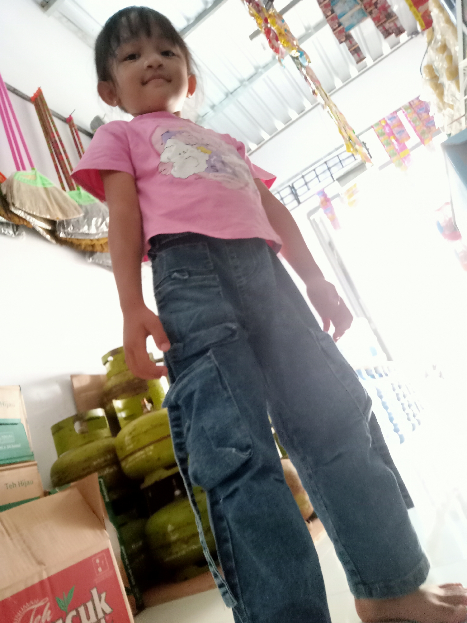 In a brightly lit interior, a young Indonesian girl stands facing the camera at a low angle. She wears a pink t-shirt with a cartoon graphic and blue denim cargo pants. Her dark hair is cut in a bob. Behind her, various items are stacked and displayed. To her left are several green propane tanks and boxes of what appears to be tea. Hanging from the ceiling are strings of colorful snacks or candies. The overall impression is of a humble, possibly domestic or small shop environment in Simo, Indonesia.