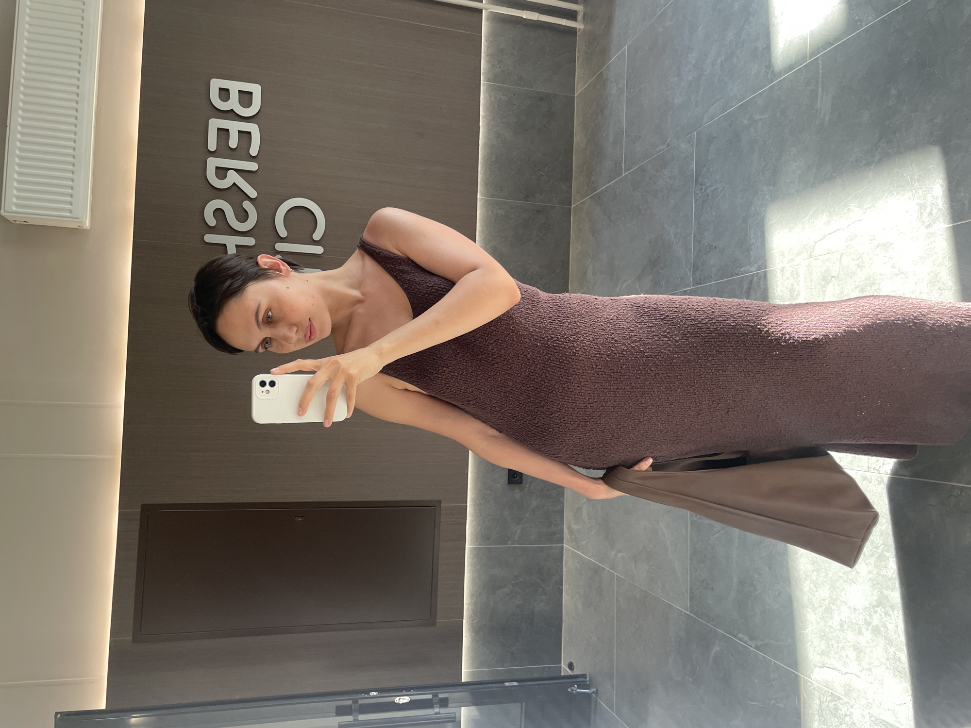 A woman is taking a selfie in what appears to be a commercial or retail setting. The background features a wall with the name "BEB2R CLINIC" partially visible, the location is most likely in the city of Izhevsk, Russia. She is wearing a brown dress, and is holding a brown leather bag in her left hand while using a white smartphone to take the photo. The floor is tiled with gray stone-like material.