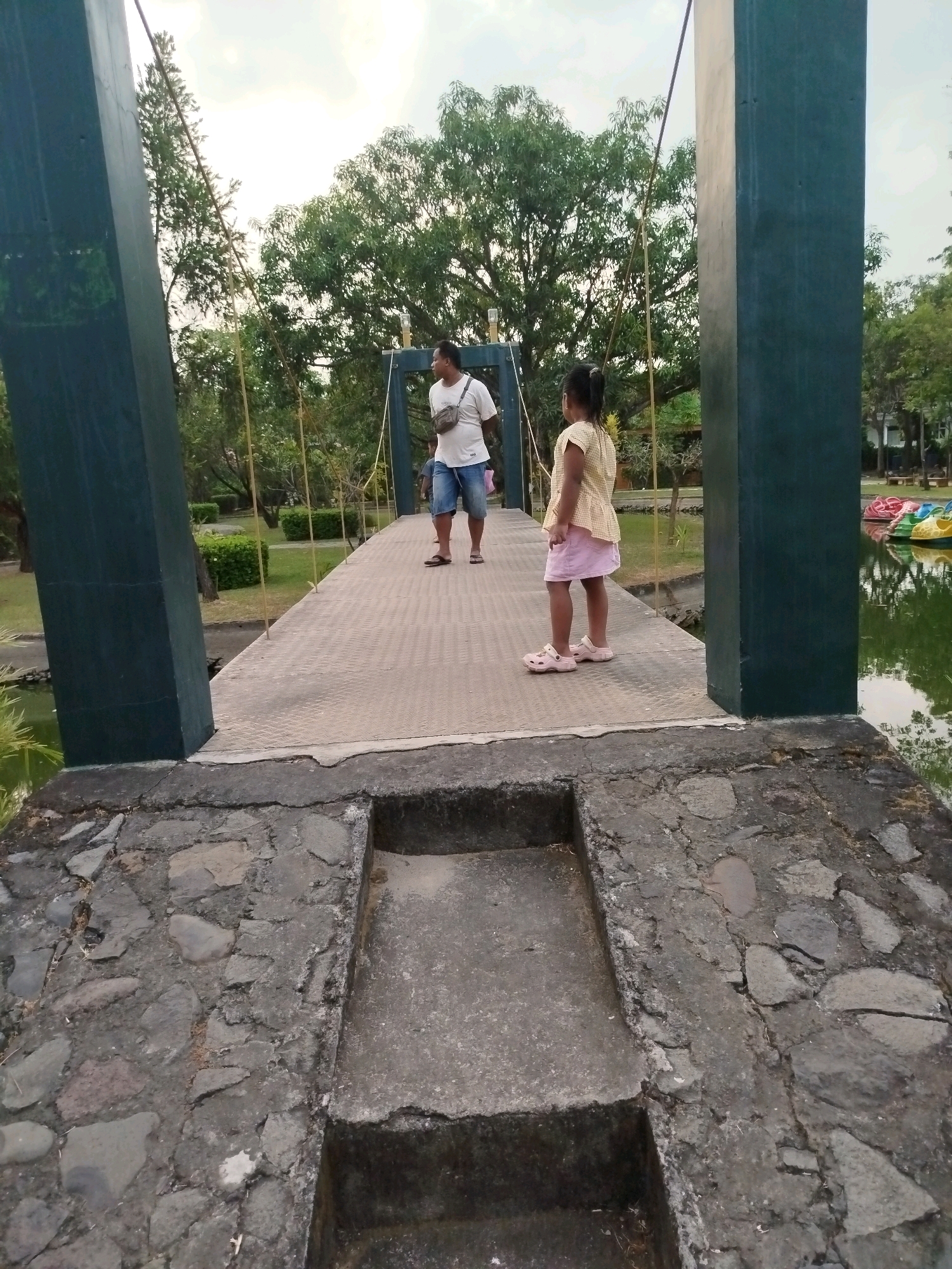 An eye-level, medium shot captures two individuals on a suspension bridge at Bugangin, Indonesia. On the left side of the bridge, a man wearing a white t-shirt and denim shorts walks away from the camera. On the right side, a young girl in a yellow checkered shirt and pink skirt faces the man. The bridge's walkway is constructed with a metallic diamond plate pattern, supported by two large, dark green concrete pillars. The bridge spans over a body of water, where several colorful paddle boats are visible on the right side. The surroundings are lush with green trees and manicured bushes, suggesting a park or recreational area. The sky is overcast with a bright, diffused light. The foreground of the image shows the weathered stone and concrete foundation of the bridge.