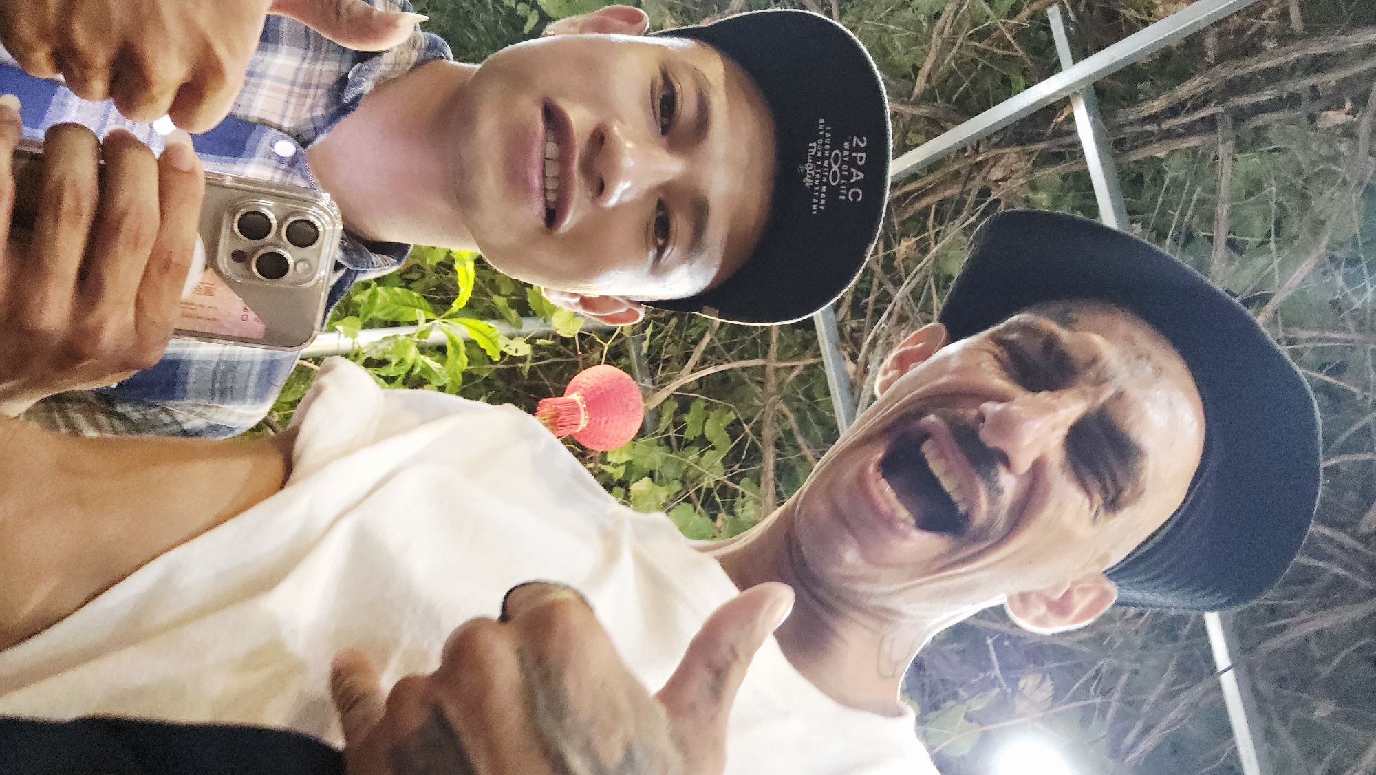 Two men are captured in a low-angle selfie, seemingly taken by the man on the left, against a backdrop of green foliage, a red lantern, and overhead metal structures, likely at night or in the evening given the artificial lighting.
The man on the left, younger, is smiling broadly at the camera. He wears a blue and white plaid shirt and a black baseball cap with white text that reads "2PAC", "WAY OF LIFE", and "DON'T LOOK DOWN ON ME ANY". He holds a smartphone with a clear case and a triple-lens camera setup (resembling an iPhone) in his left hand, positioned to take the photo.
The man on the right, older, expresses exuberant joy with a wide-open mouth, as if laughing or shouting. He sports a mustache and wears a white t-shirt and a dark, possibly wide-brimmed hat. His left hand, which has visible tattoos, is giving a thumbs-up gesture towards the camera.
The overall mood of the image is lively and jovial. The presence of the red lantern and the provided location context of Bac Nha Trang Ward, Vietnam, suggest an outdoor or semi-outdoor setting within an Asian cultural environment, possibly a restaurant, bar, or garden area.